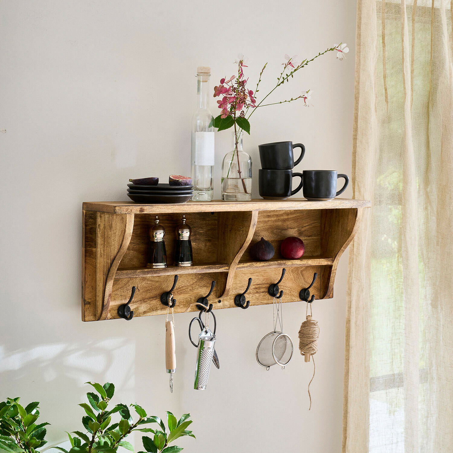 Estantería de pared Farrowby Estante de pared de madera con ganchos, tazas negras, platos y decoración floral en interior luminoso