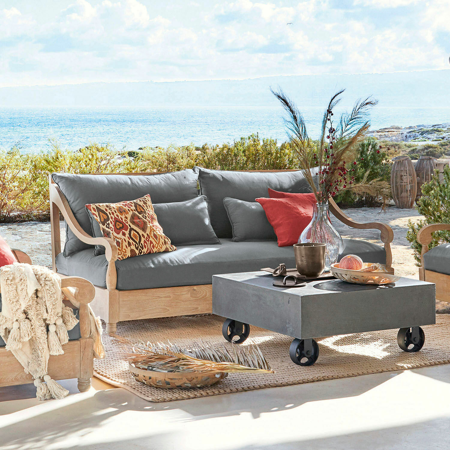 Sofá Montcassin Sofá de exterior de madera clara con cojines grises y cojines decorativos coloridos en terraza con vista al mar