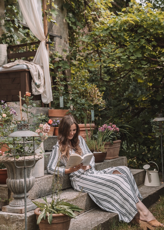Mujer con vestido a rayas leyendo un libro en escalones de piedra rodeada de macetas y flores en jardín