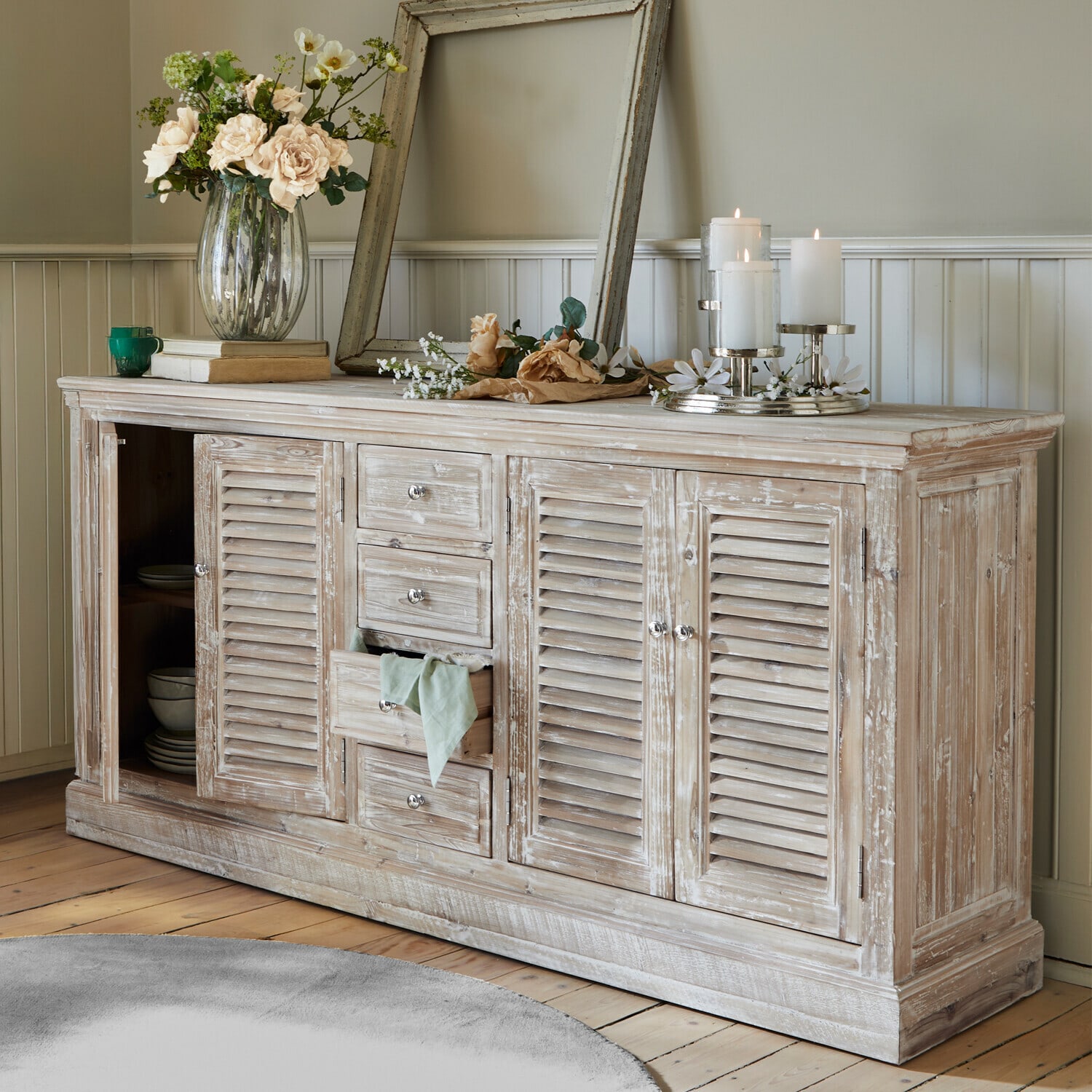 Aparador rústico de madera con acabado blanco desgastado, decoraciones florales y velas en salón