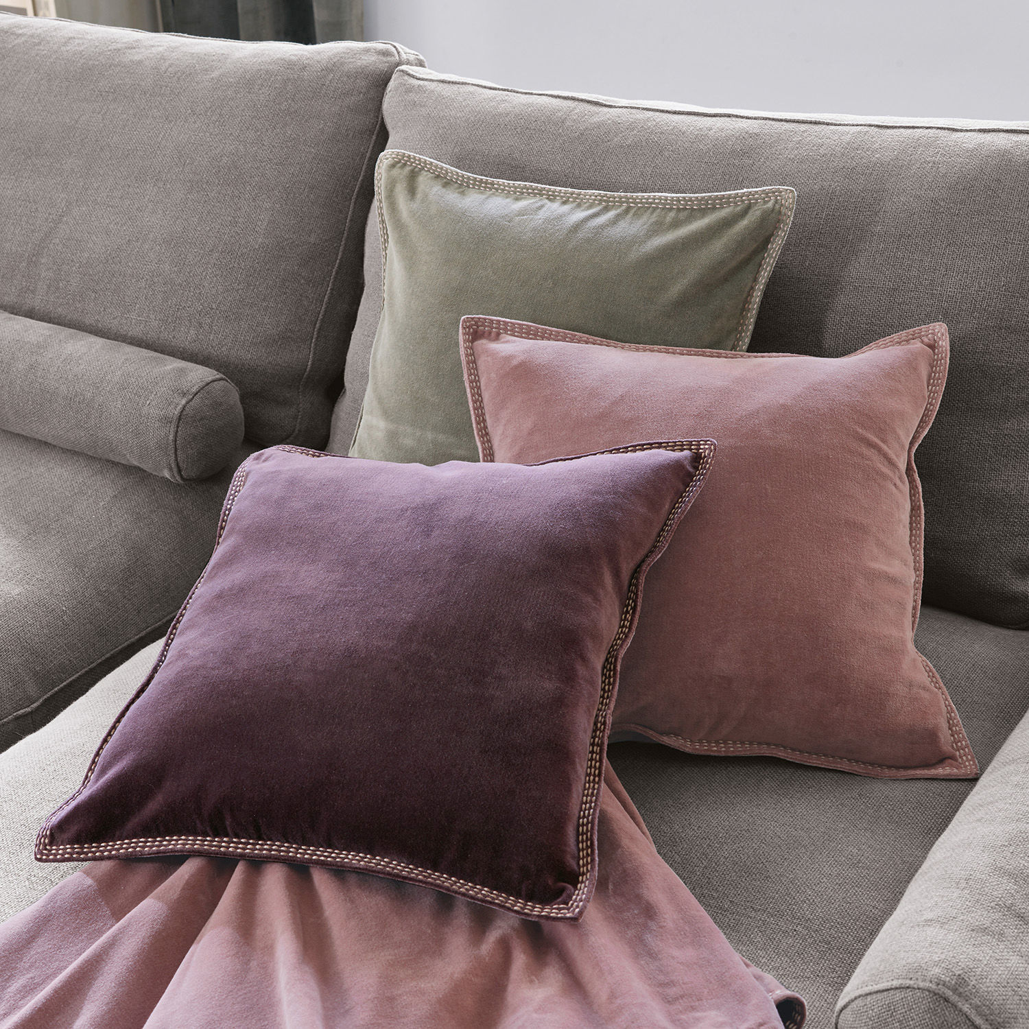 Set de 3 cojines Allonnes Conjunto de tres cojines de terciopelo en tonos violeta, rosa y verde sobre un sofá gris