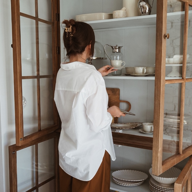 Mujer con camisa blanca cogiendo taza de porcelana en vitrina de madera con vajilla