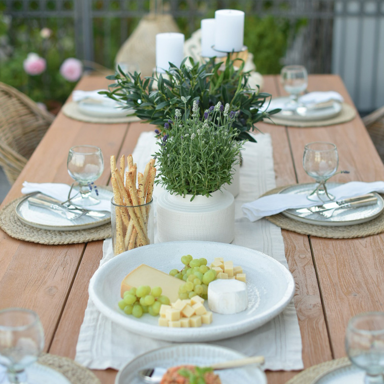 Mesa de madera con vajilla blanca, quesos, uvas verdes y decoración vegetal para comida al aire libre