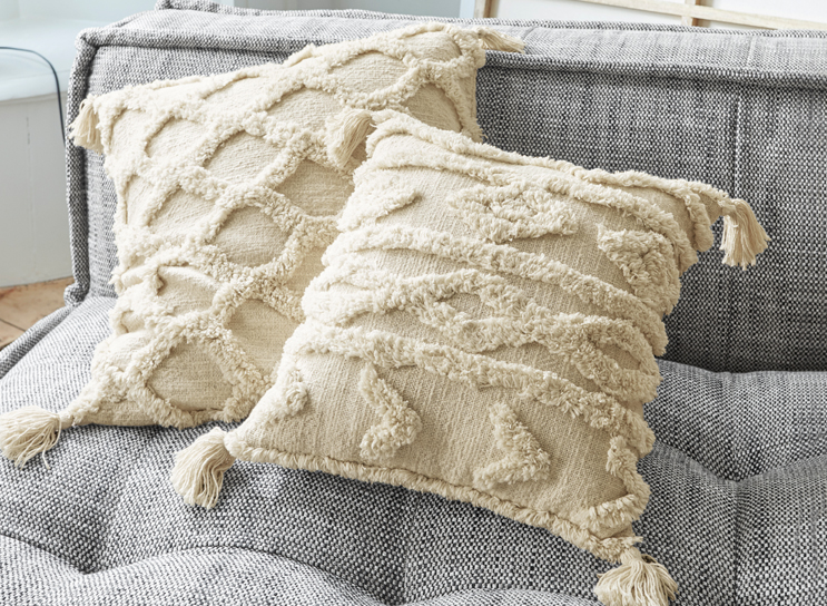 Cojines decorativos beige con flecos y patrón geométrico sobre sofá gris de tela