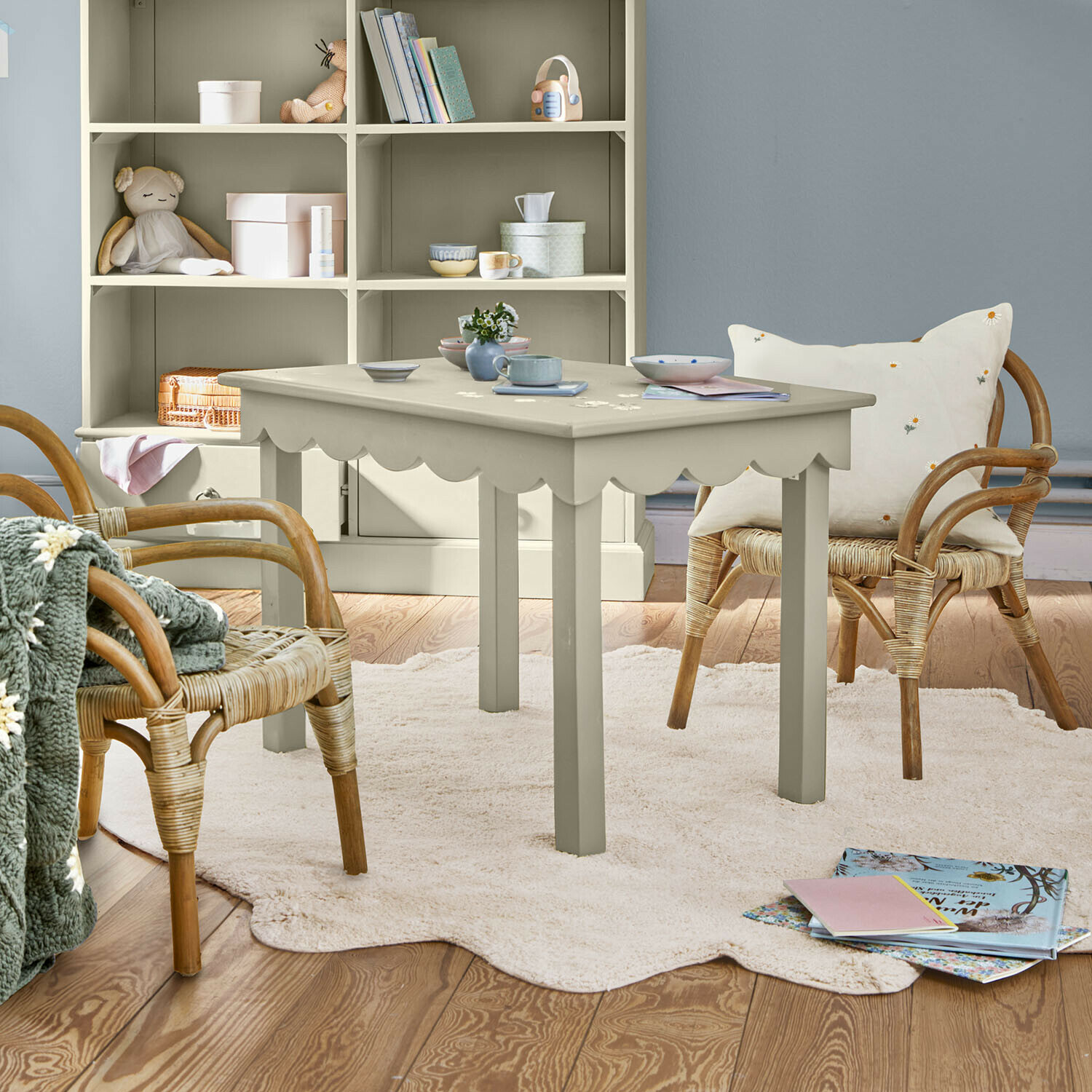Mesa de madera gris con borde ondulado sobre alfombra clara en habitación infantil acogedora con sillas de mimbre
