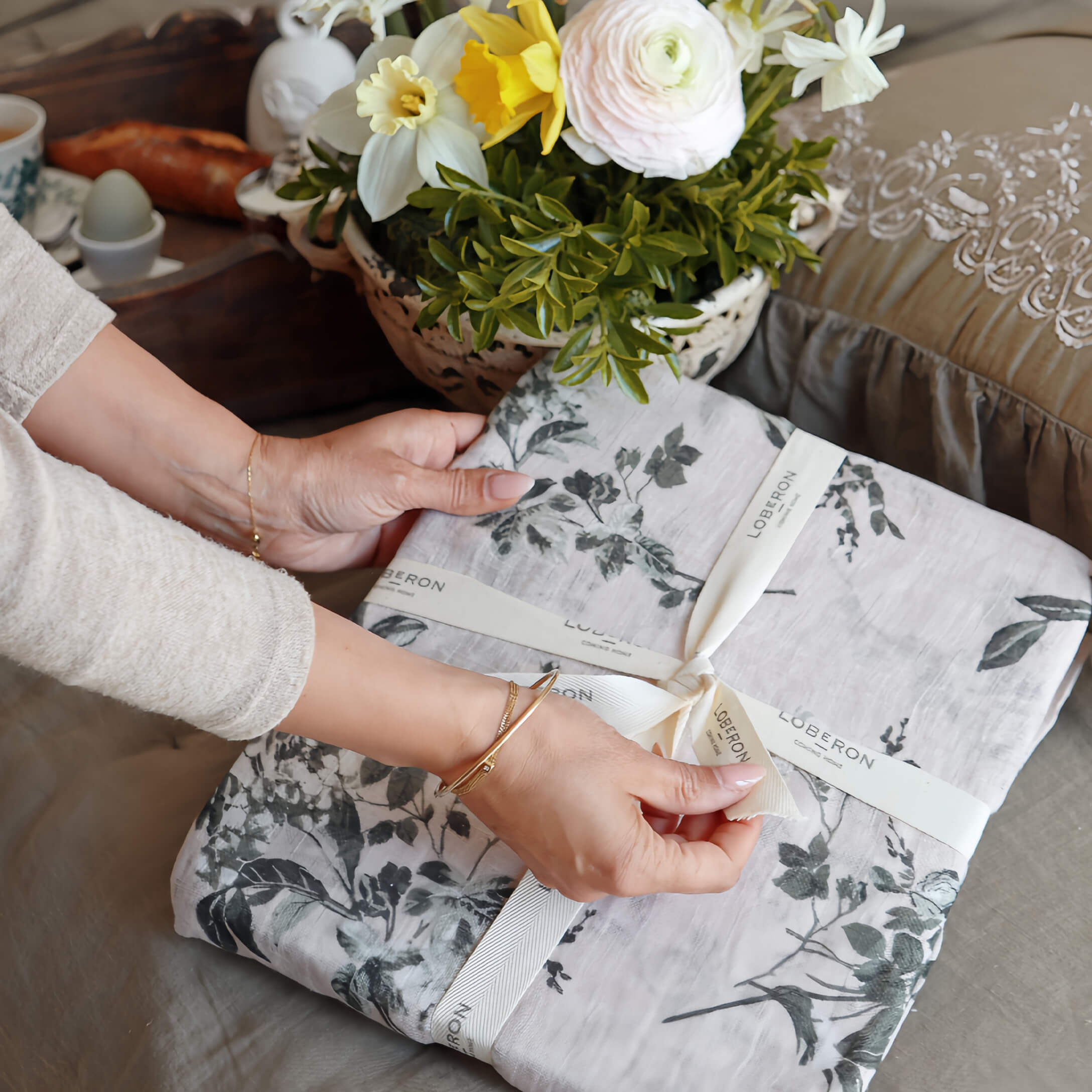 Manos desenvolviendo ropa de cama de lino gris con diseño vegetal, flores decorativas al fondo
