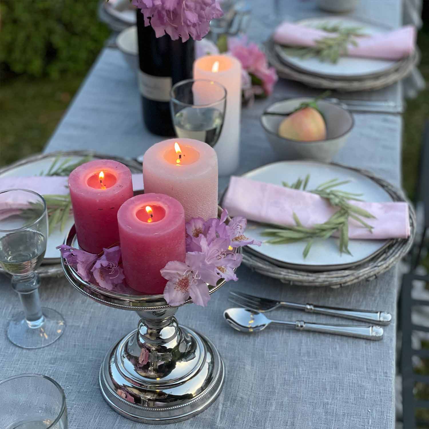 Candelabro plateado con tres velas encendidas rosas y blancas sobre mantel gris con flores decorativas