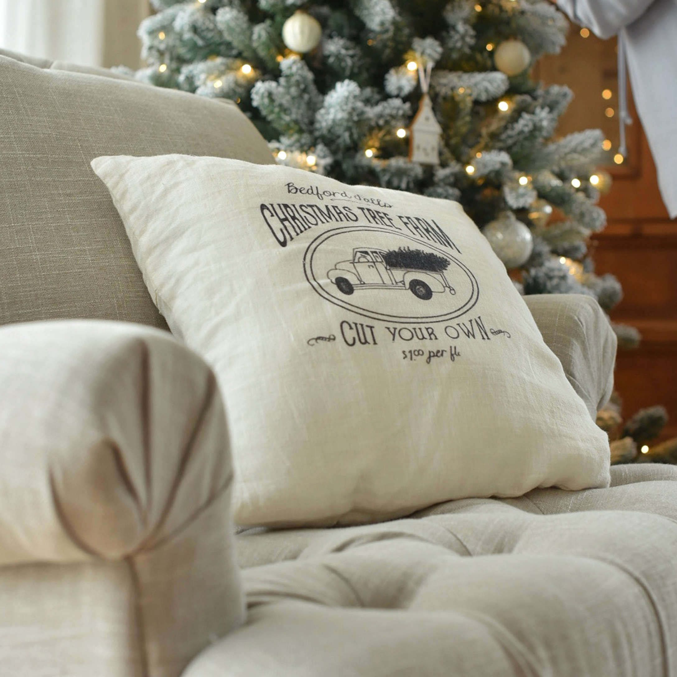 Cojín beige de lino con estampado de camión navideño en sofá junto a árbol de Navidad decorado