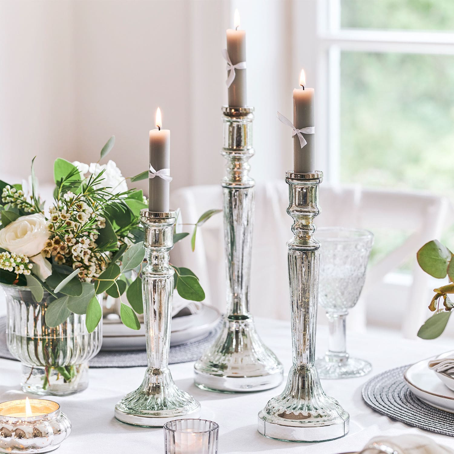 Set de 3 candeleros Riley Tres candelabros plateados con velas grises sobre mesa blanca con decoración floral y cristal