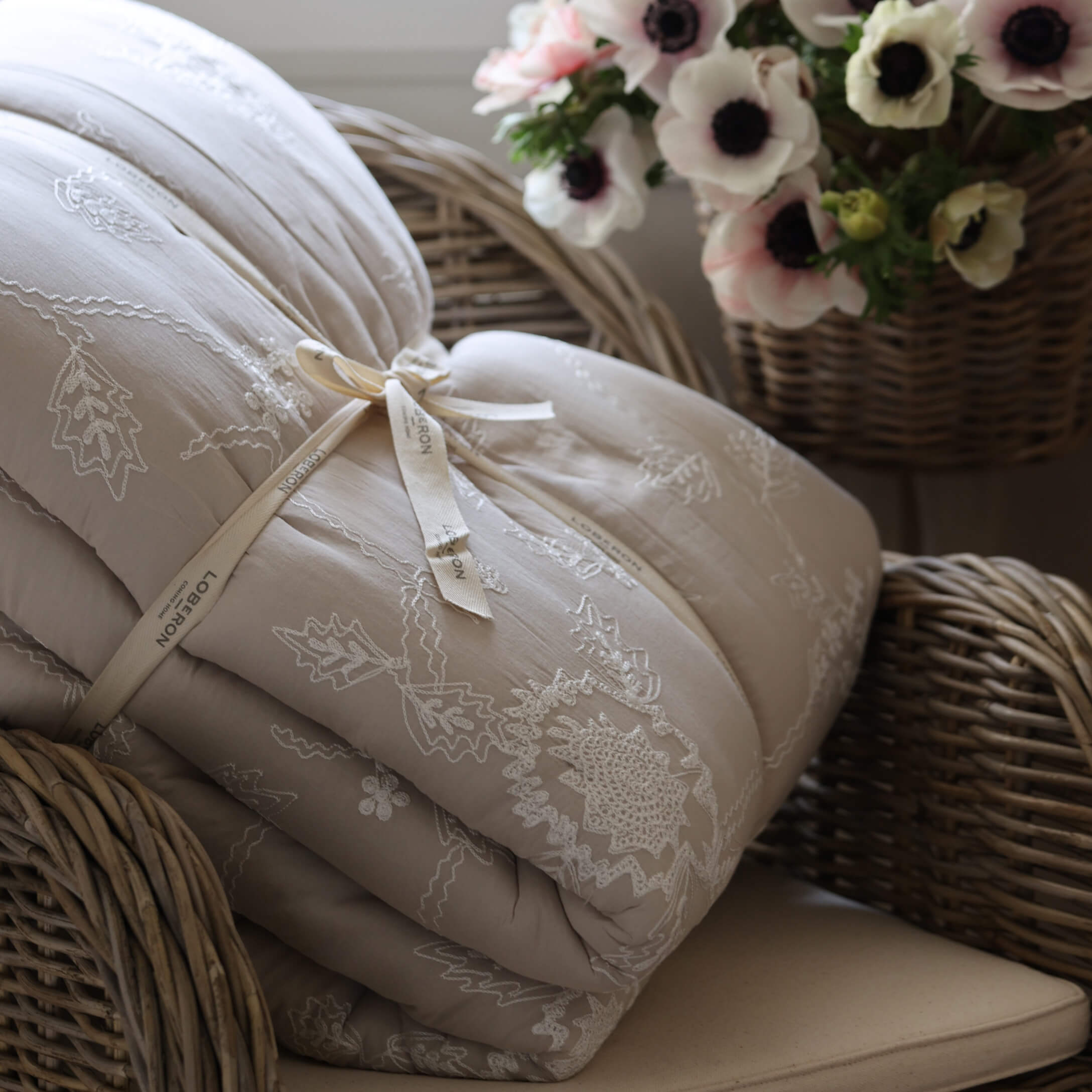 Colcha beige acolchada con bordado sobre sillón de mimbre junto a cesta con flores, decoración hogareña