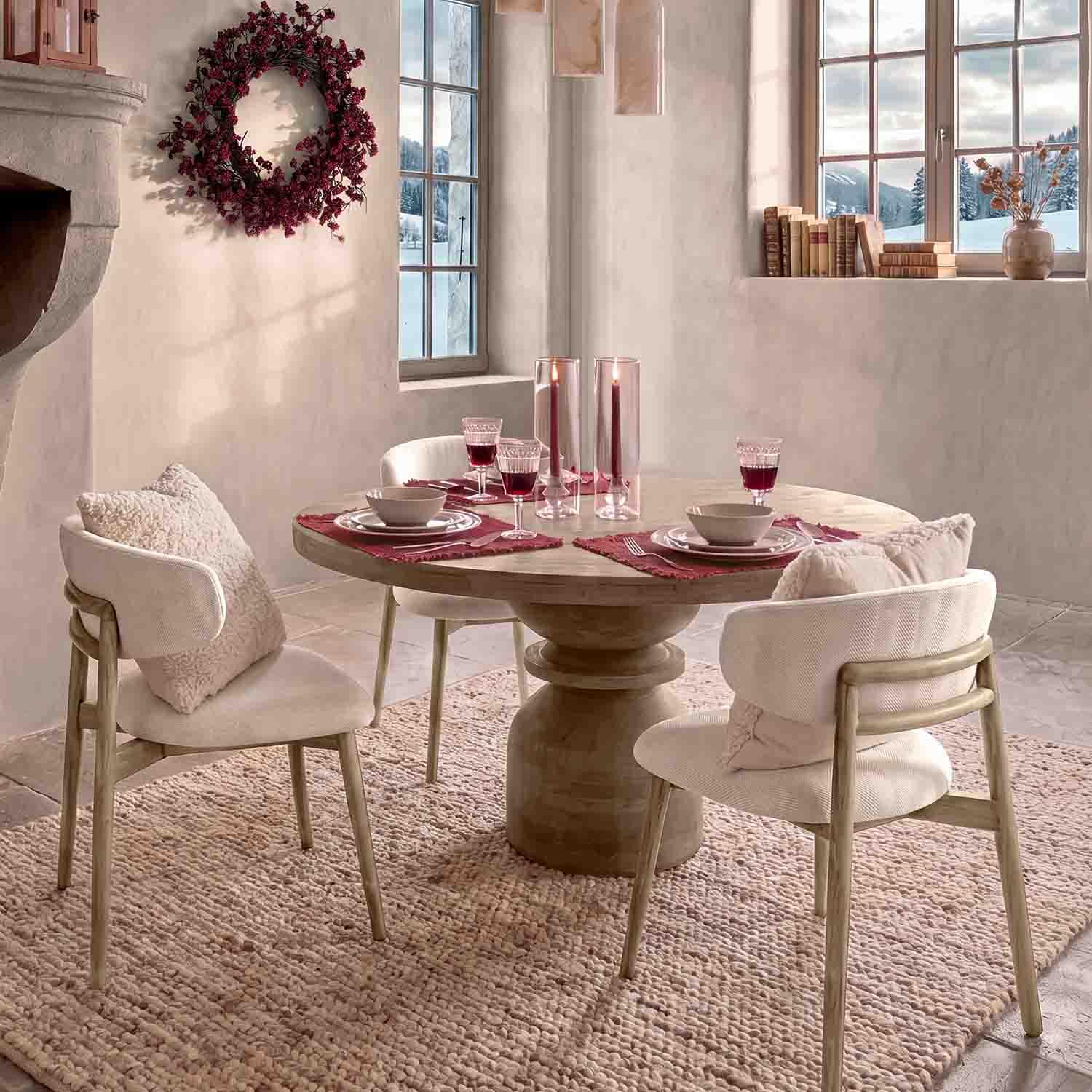 Sillas claras con cojines alrededor de mesa redonda de madera con velas rojas y copas de vino en ambiente acogedor