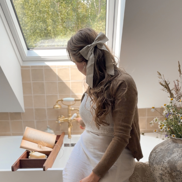 Mujer con lazo de lino beige en el cabello sentada junto a una bañera con bandeja de madera y libro, luz natural