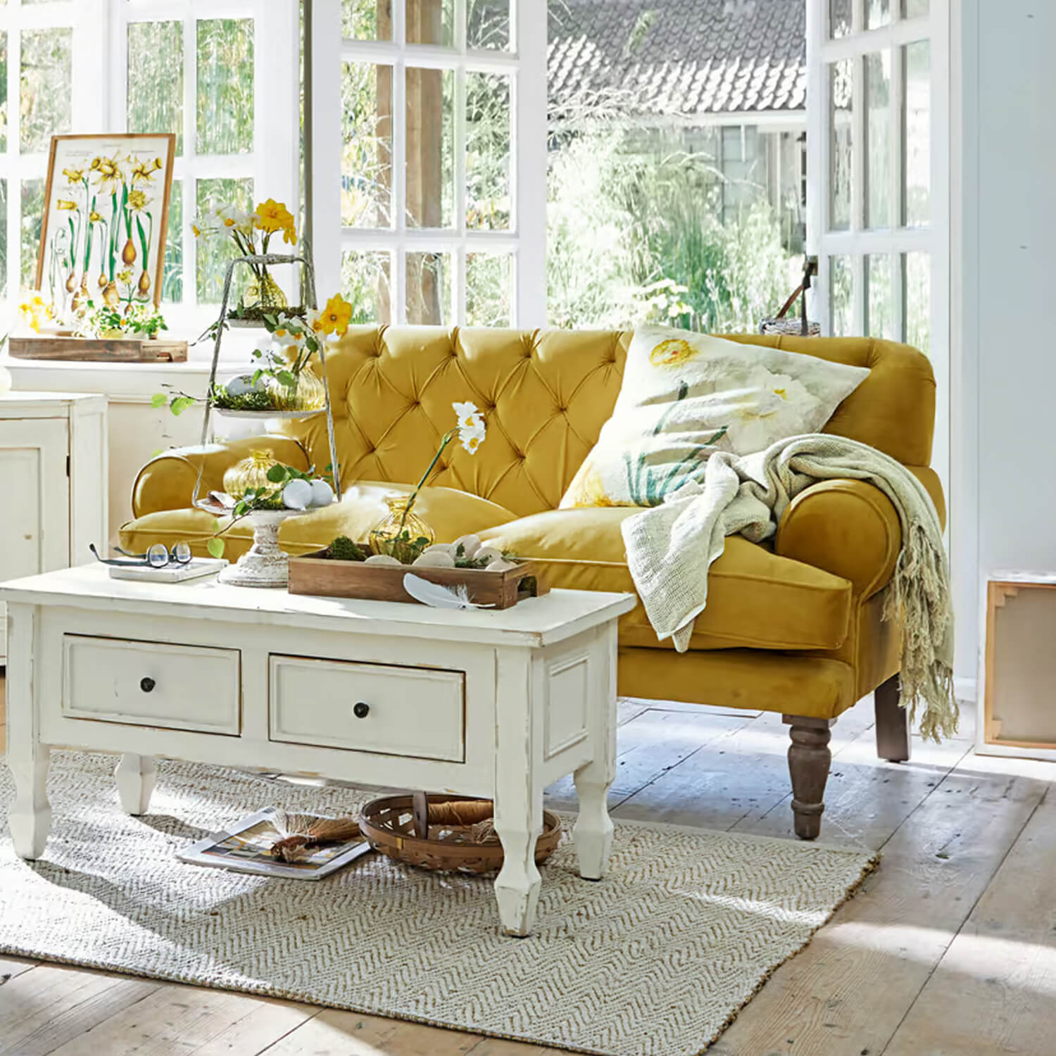 Sofá amarillo de terciopelo con respaldo capitoné en salón luminoso con mesa de centro blanca y decoración floral