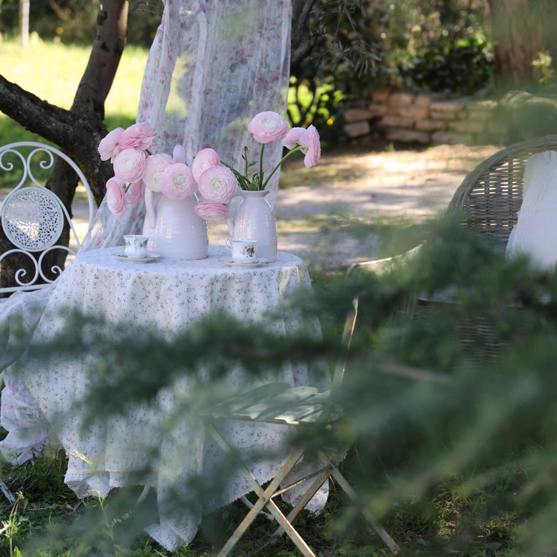 Mesa de jardín blanca con mantel floral y jarras blancas con flores rosas, ambiente romántico