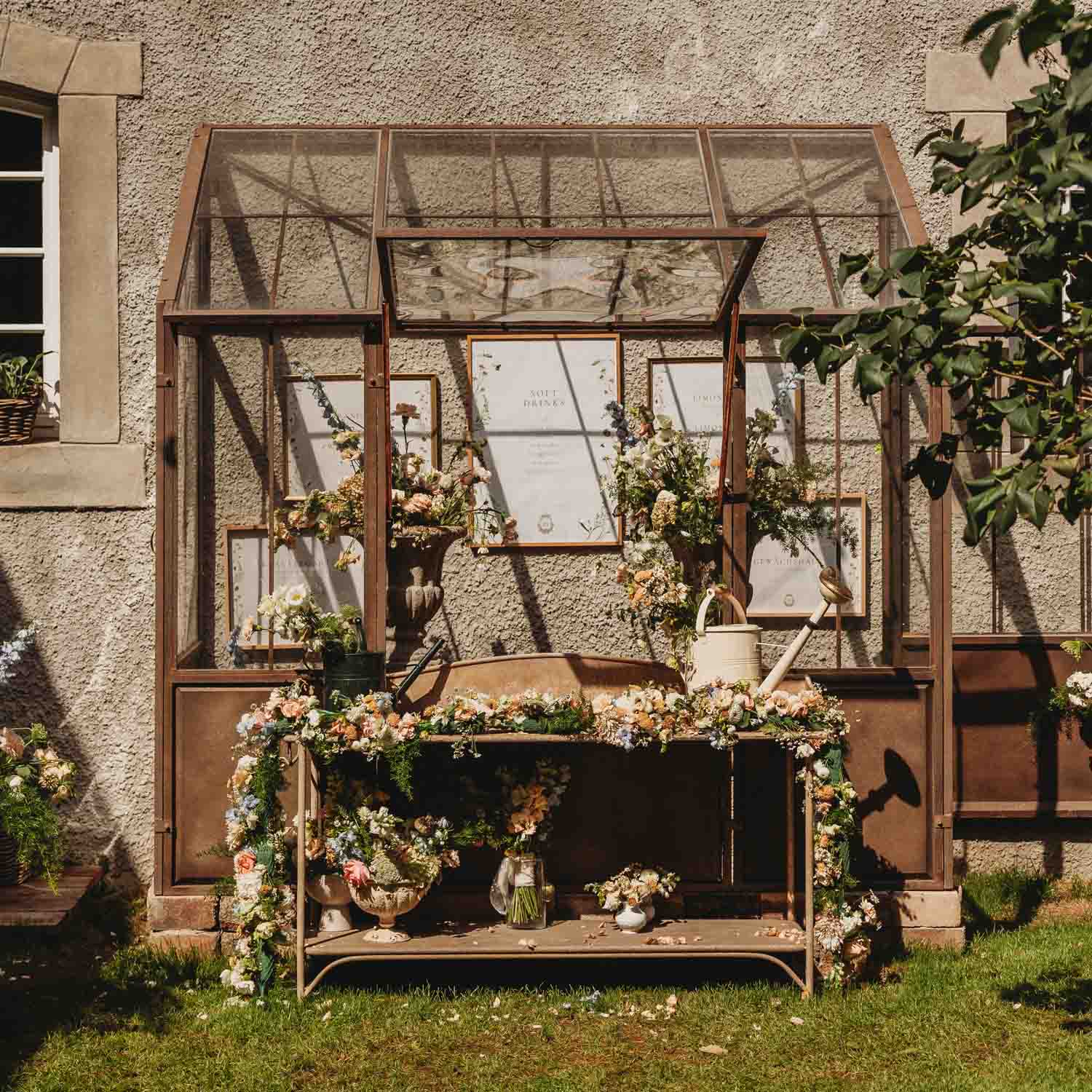 Banco de jardín de metal rústico decorado con flores y regadera frente a pared texturizada con ventana