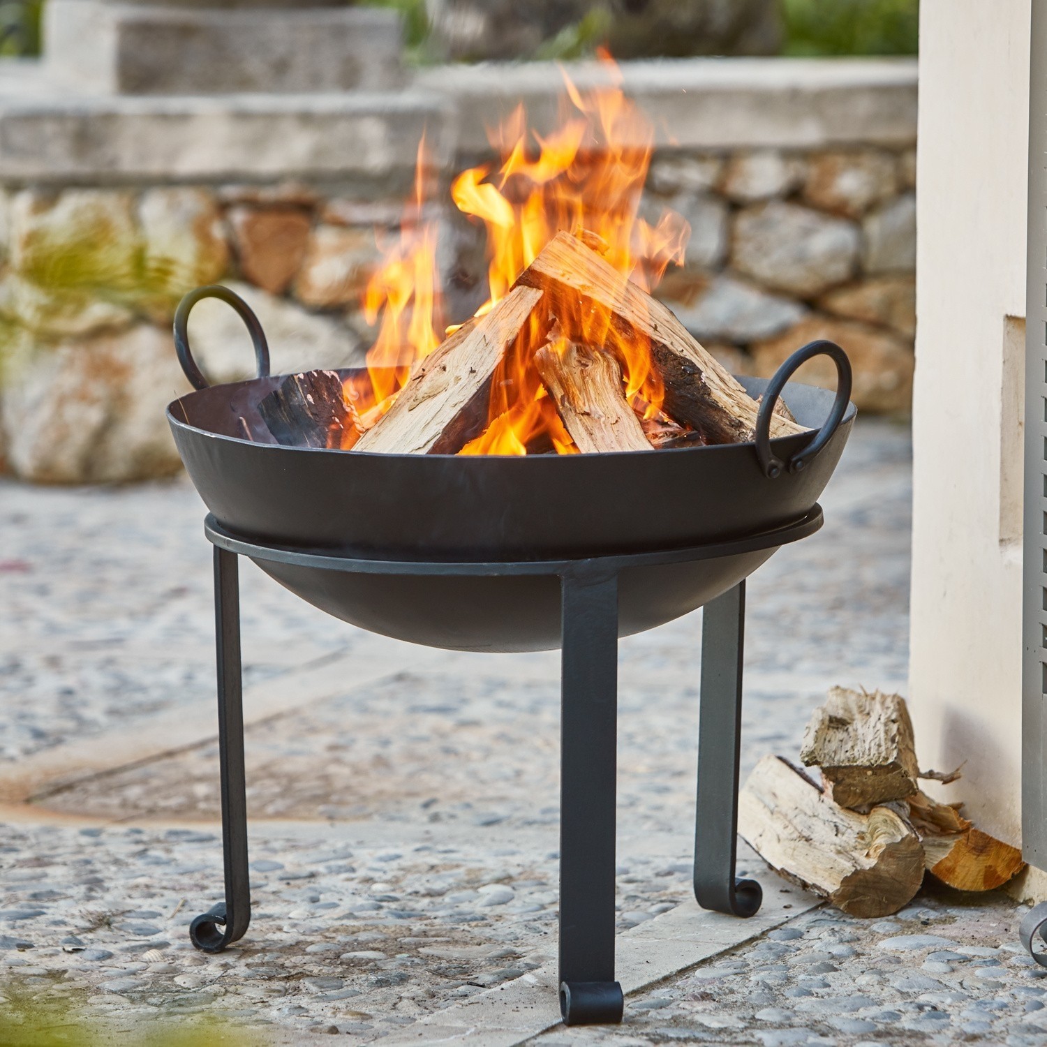 Brasero Harrogate Brasero metálico negro con leña ardiendo sobre suelo de piedra en exterior