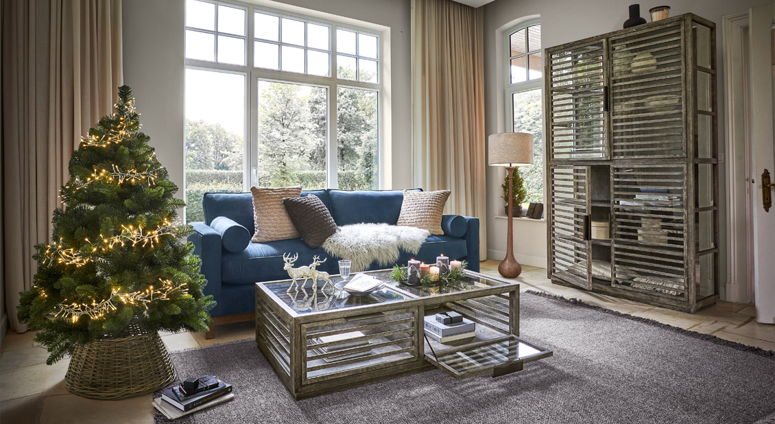 Salón con sofá azul, mesa de madera y armario, árbol de Navidad iluminado en cesta de mimbre