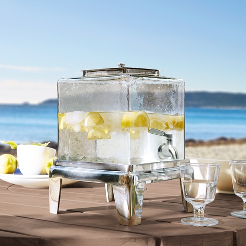 Dispensador de bebidas de vidrio con base metálica, lleno de agua con limón, ideal para fiestas de verano