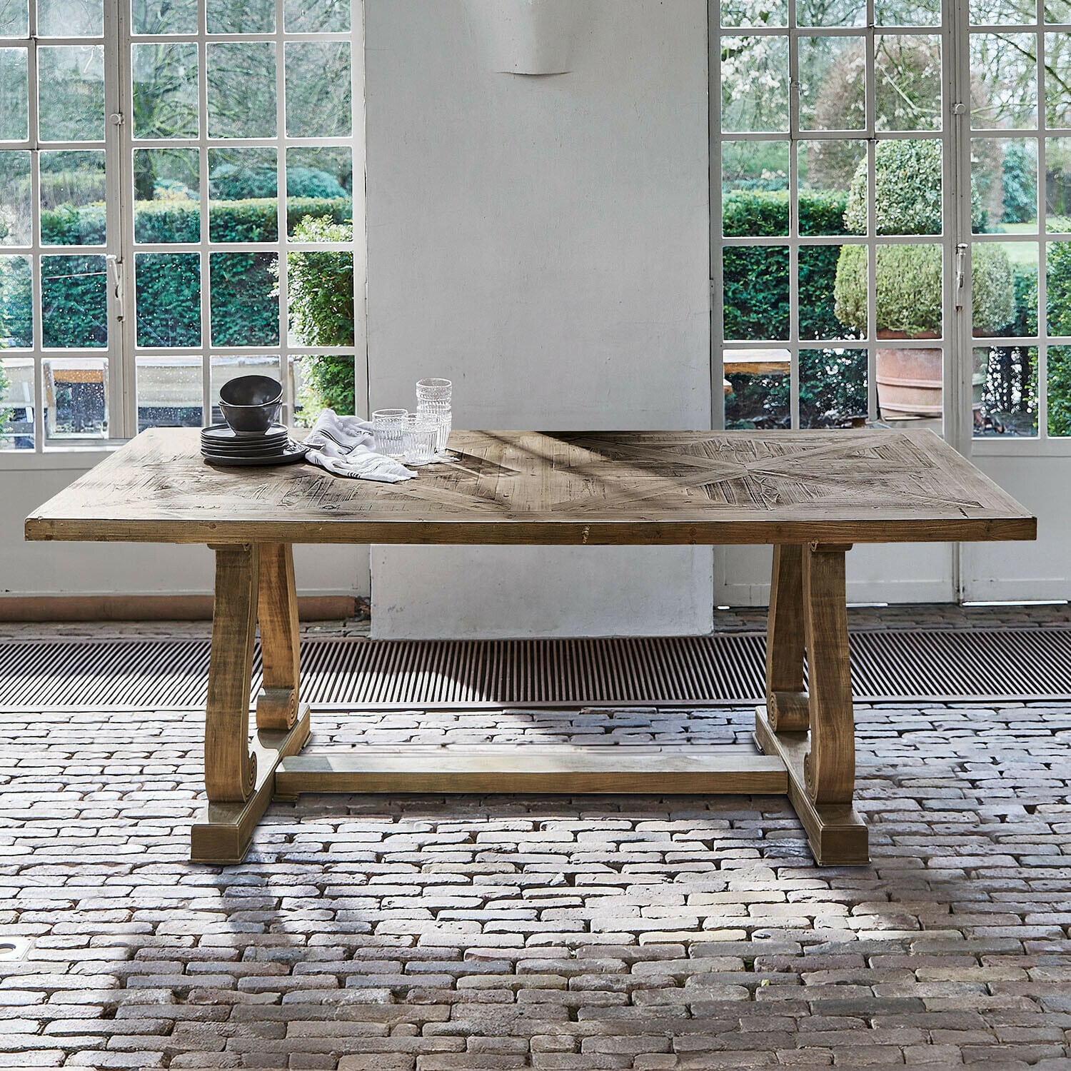 Mesa Campbelltown Mesa de madera en color natural con tablero decorativo frente a grandes ventanas y suelo de ladrillo