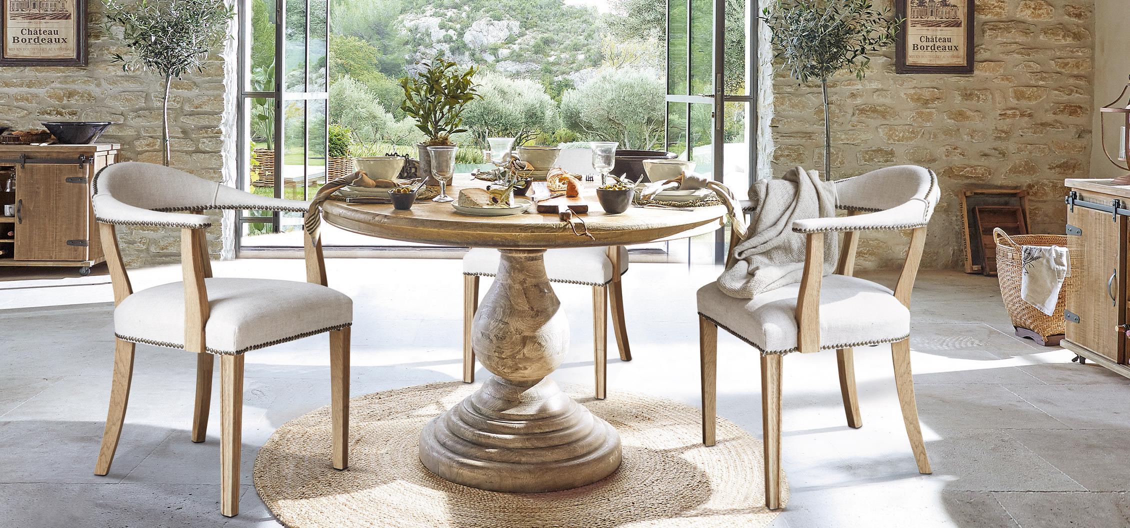 Sillas de madera clara con tapicería beige alrededor de mesa redonda en comedor rústico con vista al jardín