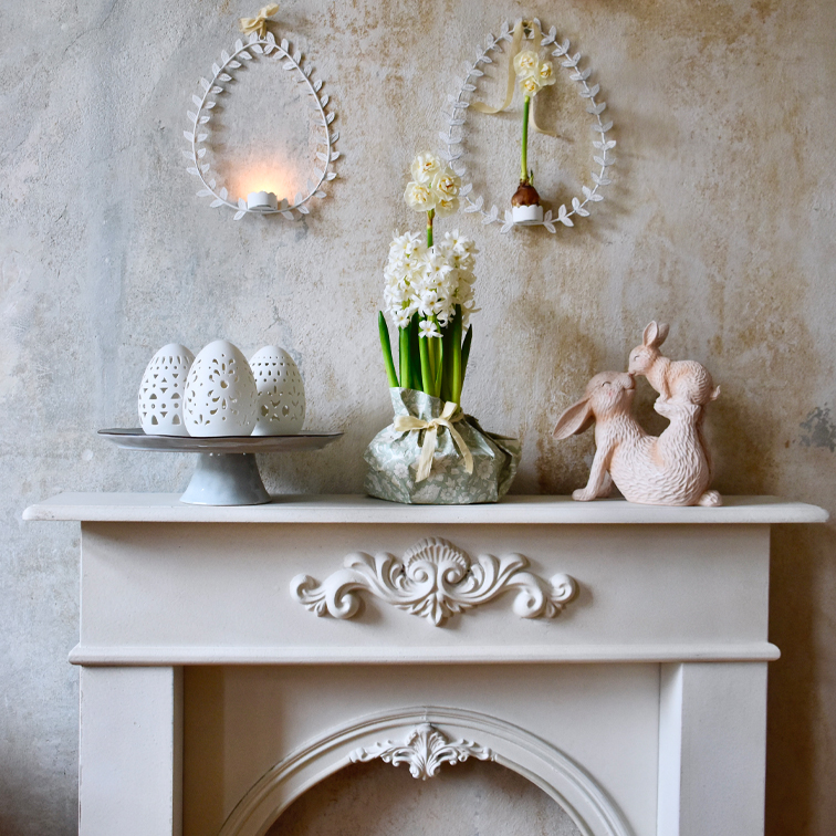 Decoración de Pascua con huevos de cerámica blanca, flores y figuras de conejos sobre una chimenea
