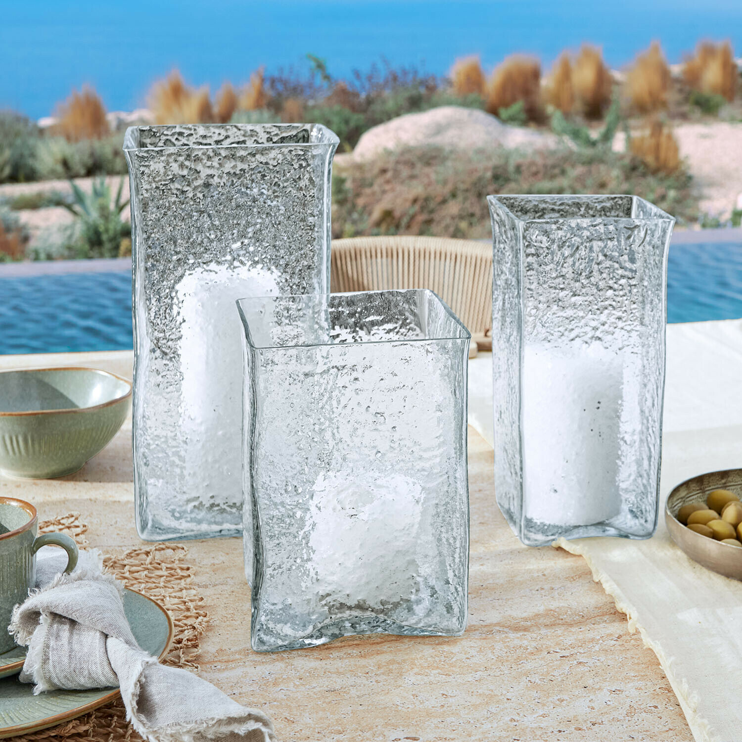 Set de 3 portavelas Xarlac Tres faroles de vidrio transparente con textura y velas blancas sobre mesa exterior junto a piscina