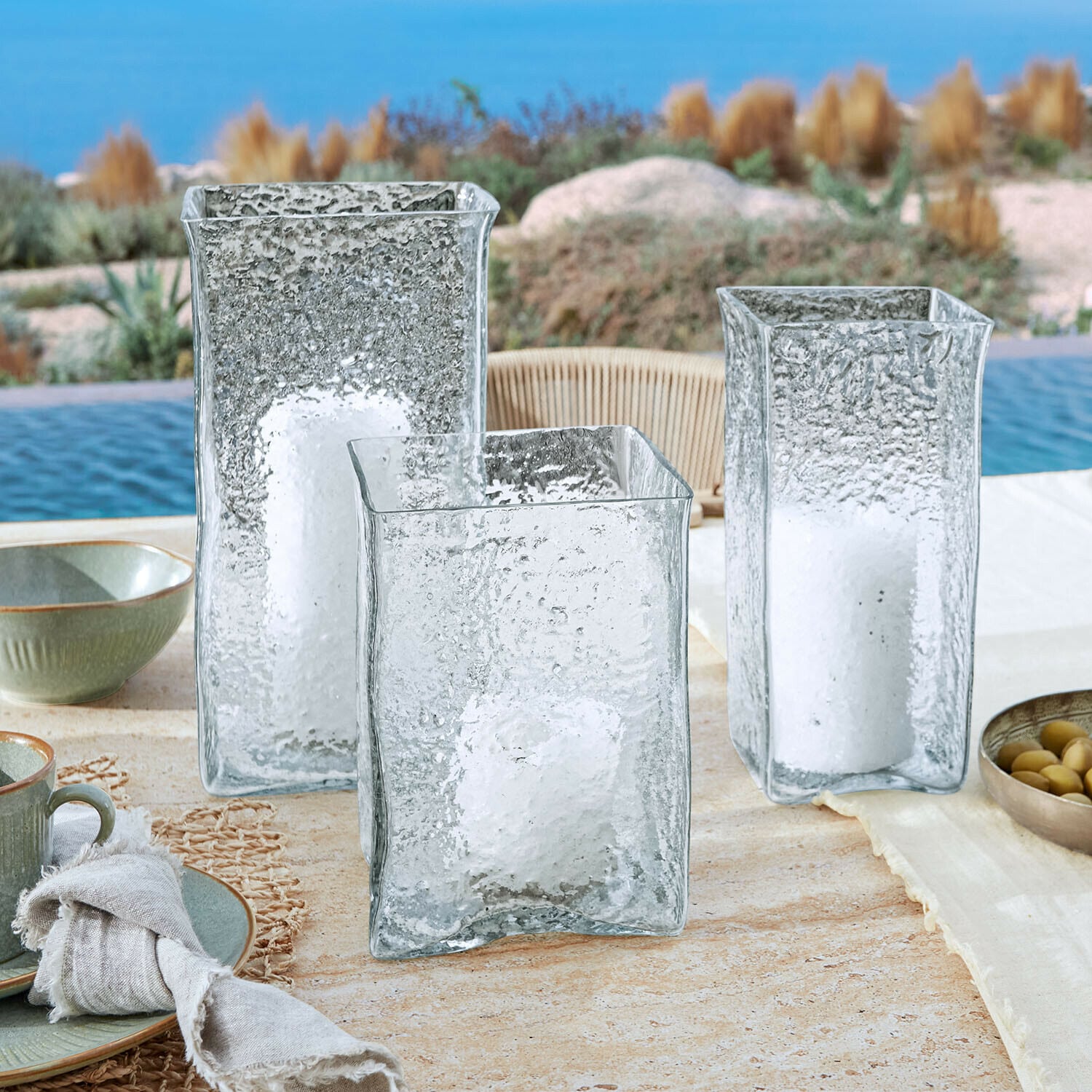 Set de 3 portavelas Xarlac Tres faroles de vidrio transparente con textura y velas blancas sobre mesa exterior junto a piscina