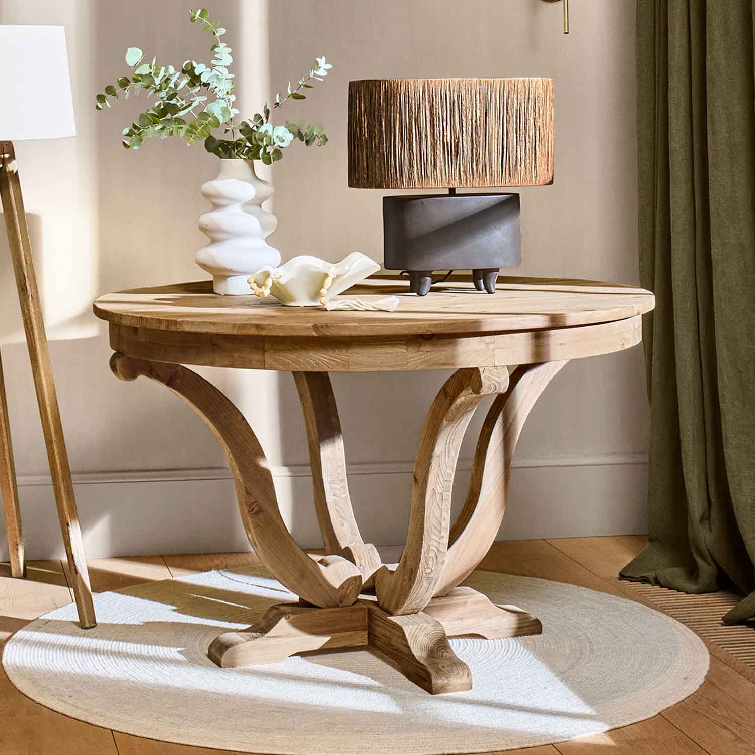 Mesa redonda de madera natural con lámpara y jarrón decorativo sobre alfombra clara en sala acogedora
