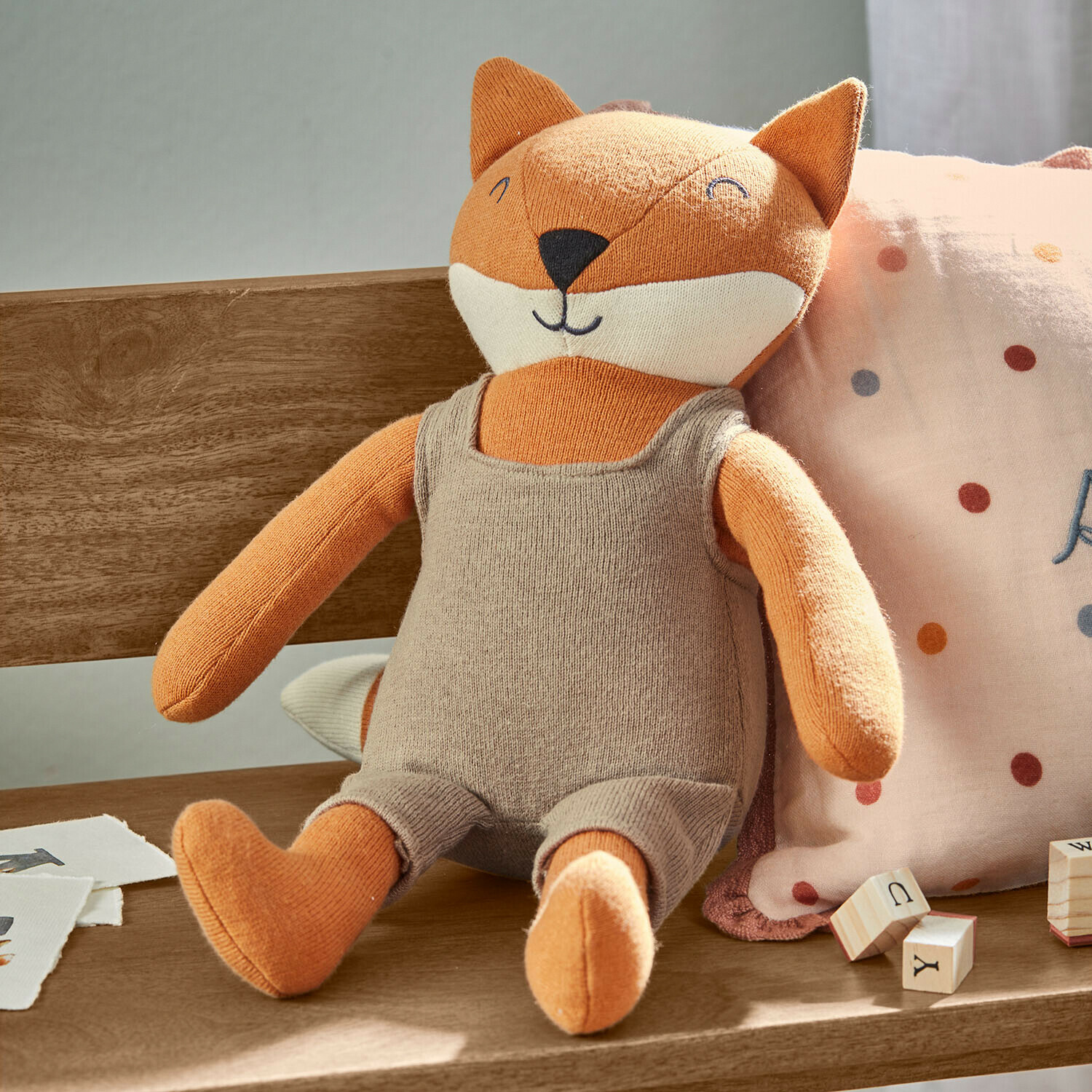 Peluche de zorro naranja con ropa beige sentado en banco de madera junto a bloques de aprendizaje