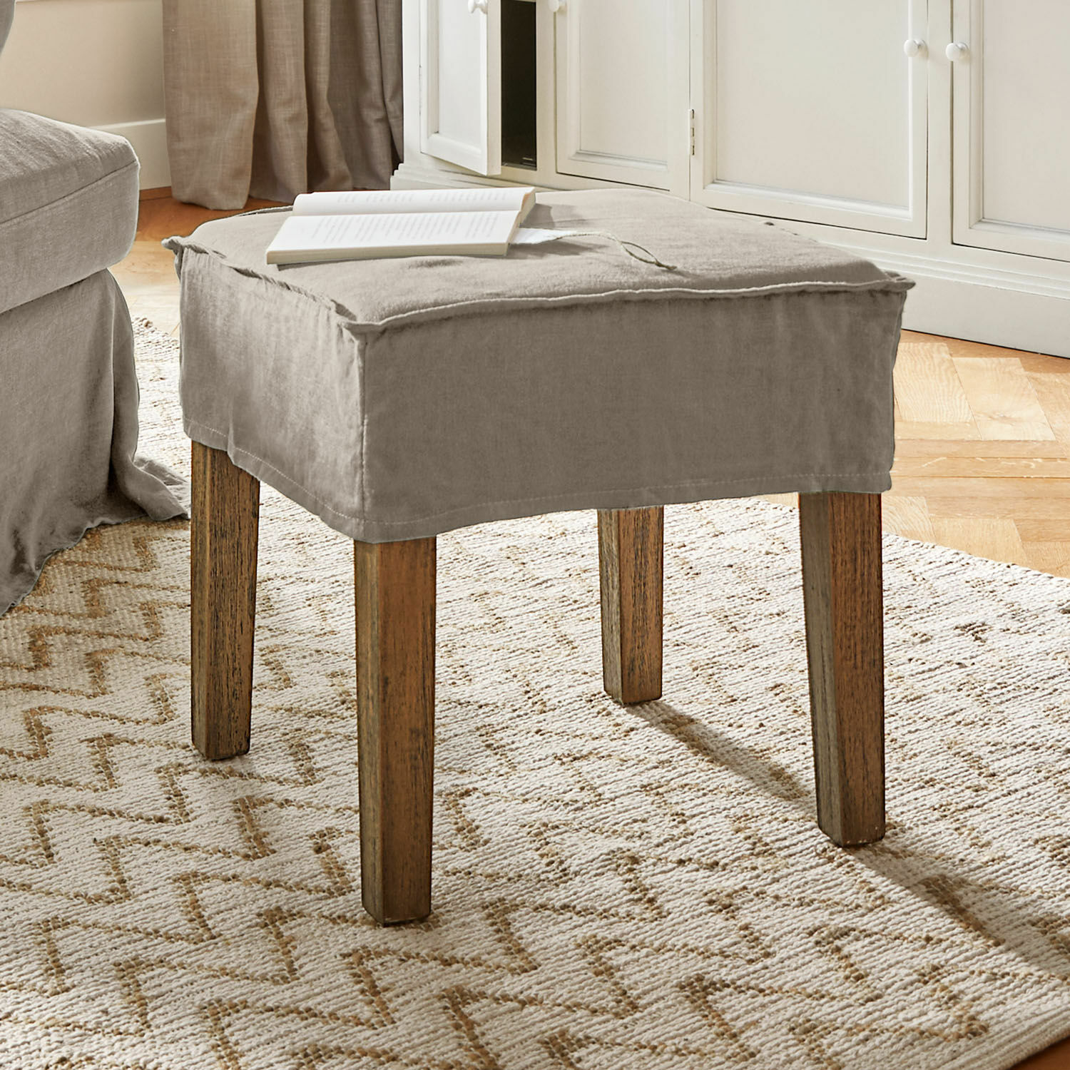Taburete Vionnique Puf beige con funda de algodón y patas de madera sobre alfombra clara en sala de estar