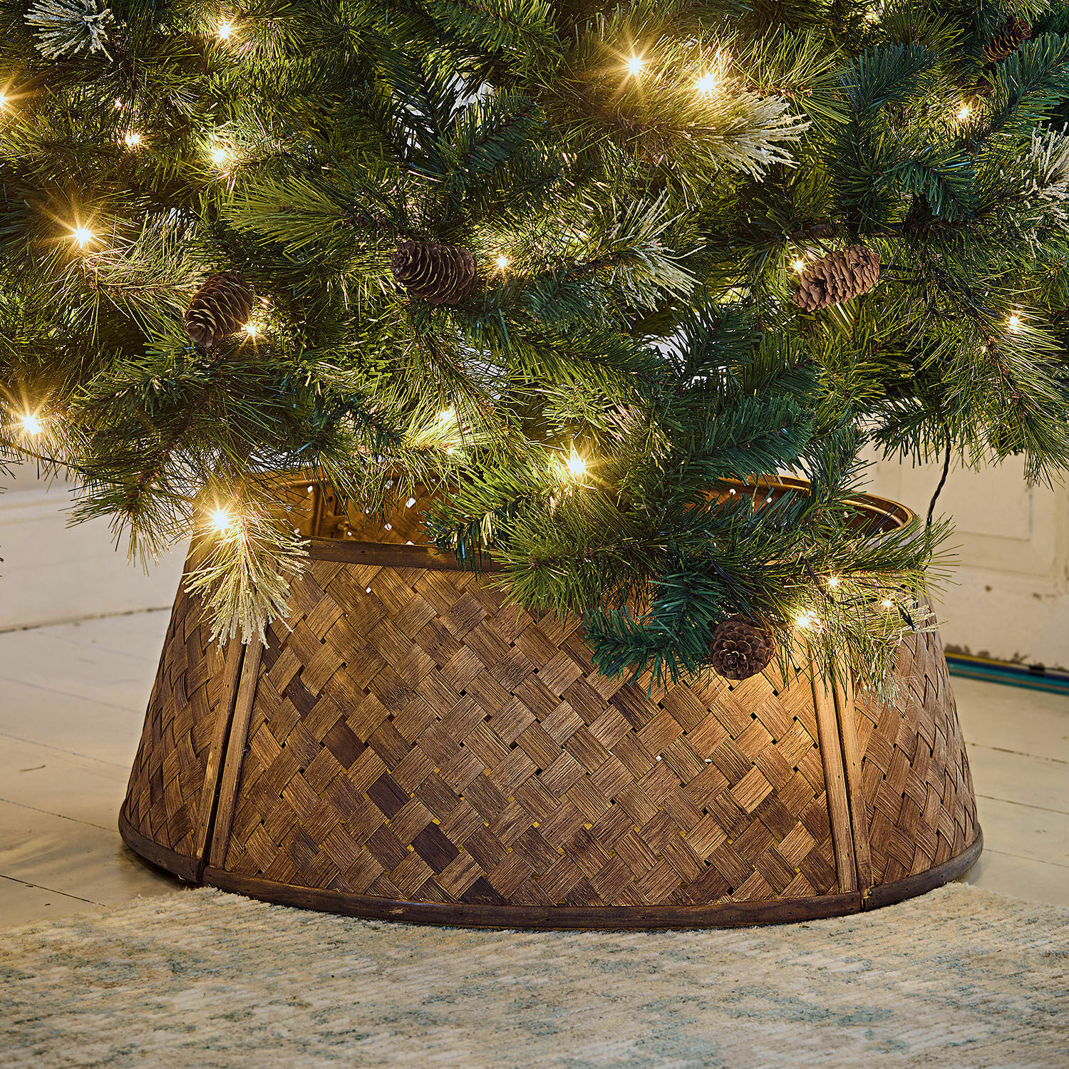 Cesta para árbol de Navidad Eden Soporte para árbol de Navidad de madera trenzada marrón bajo árbol verde con luces y piñas