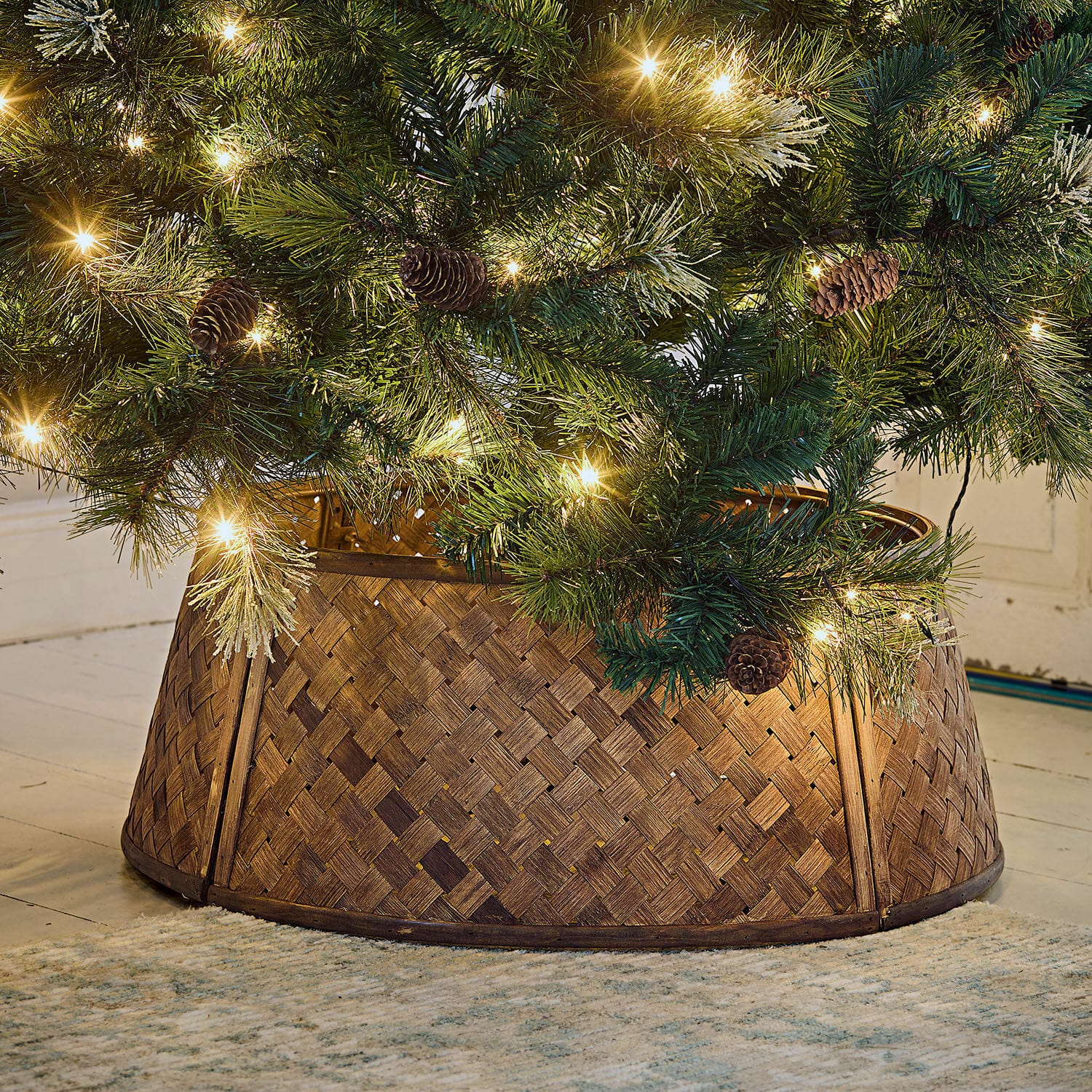 Cesta para árbol de Navidad Eden Soporte para árbol de Navidad de madera trenzada marrón bajo árbol verde con luces y piñas