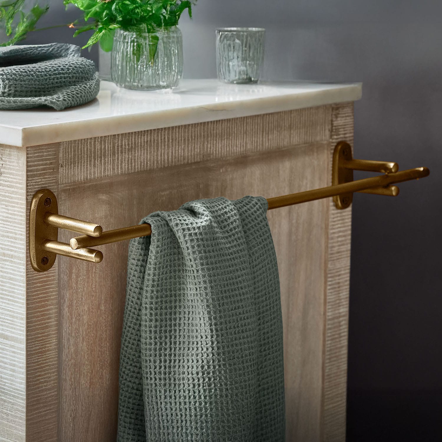 Toallero metálico dorado con toalla verde de textura waffle sobre mueble de madera con encimera de mármol