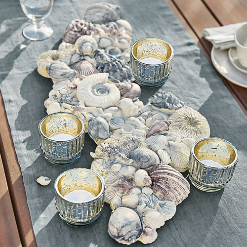 Camino de mesa decorativo con conchas grises y cuatro portavelas de vidrio dorado sobre mesa de madera