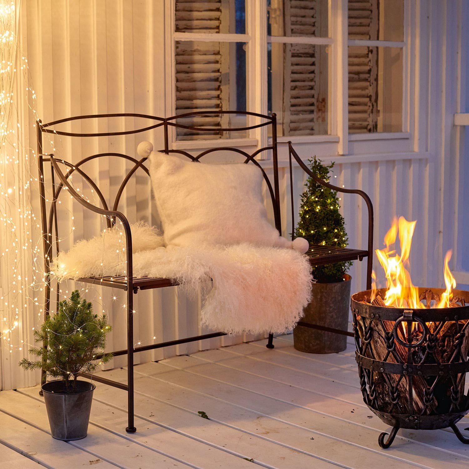 Banco Jablines Banco metálico con cojín blanco y manta de piel junto a una hoguera en terraza con luces decorativas