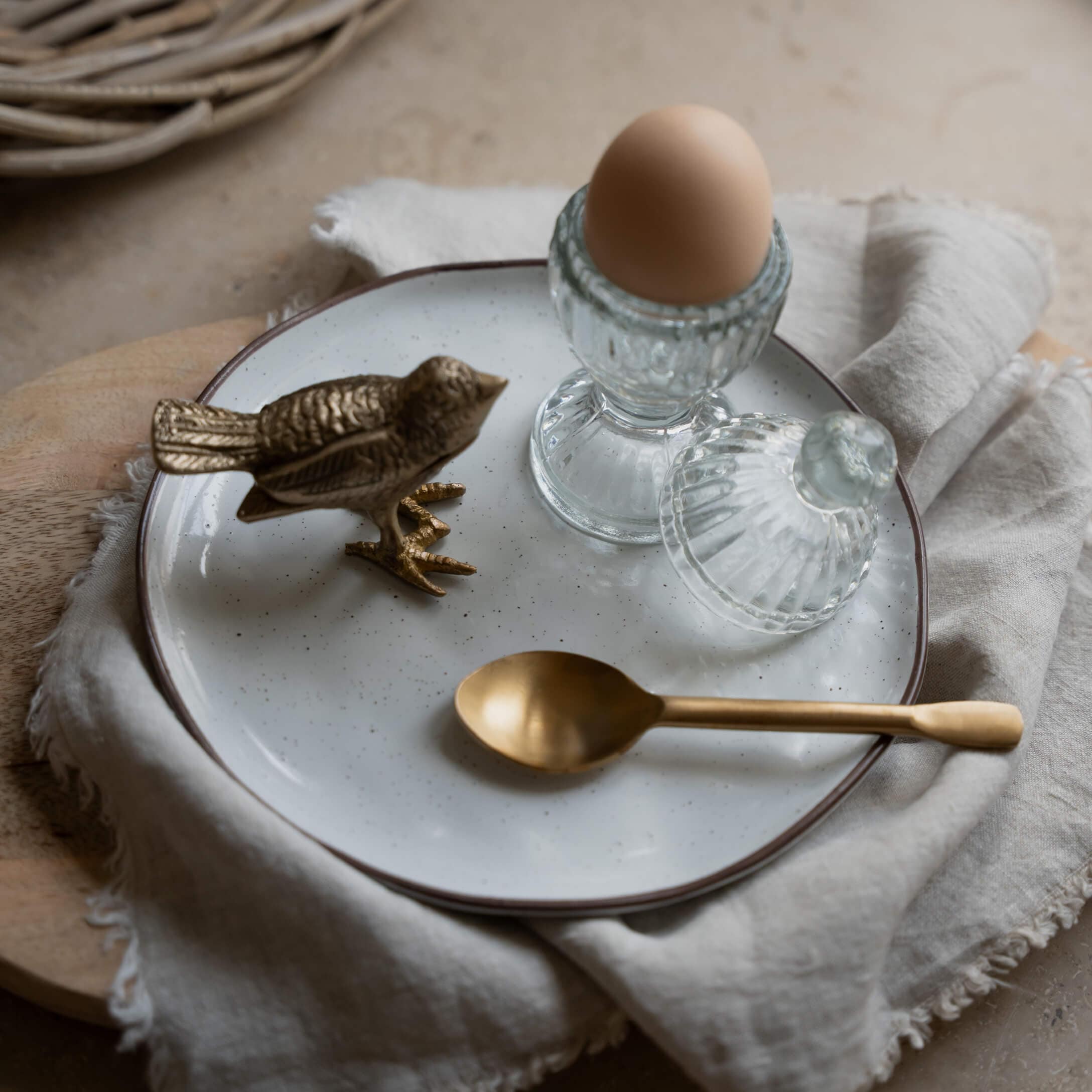 Soporte de huevo de cristal con tapa y huevo marrón sobre plato blanco con cuchara de latón y figura de pájaro