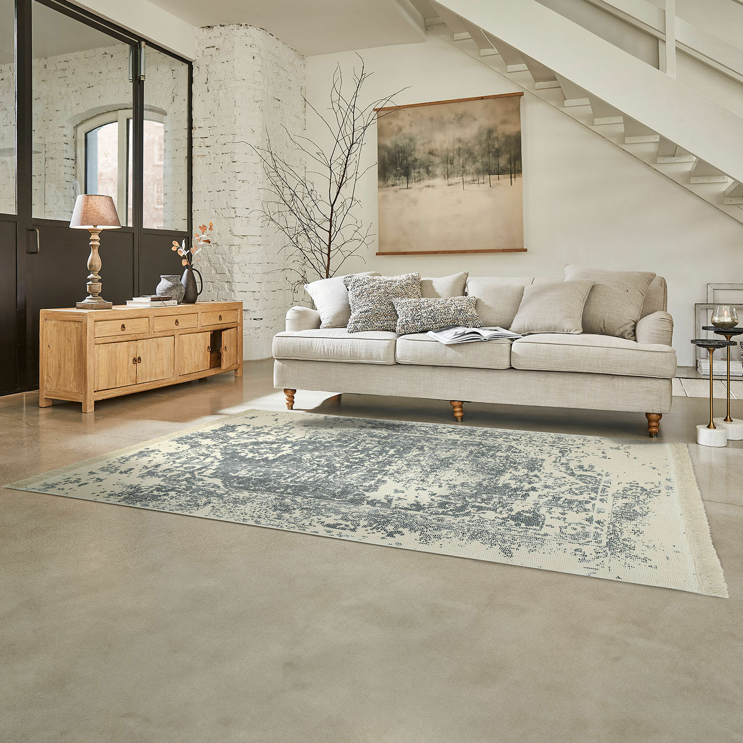 Sofá beige con cojines sobre alfombra gris en sala luminosa con cómoda de madera y lámpara
