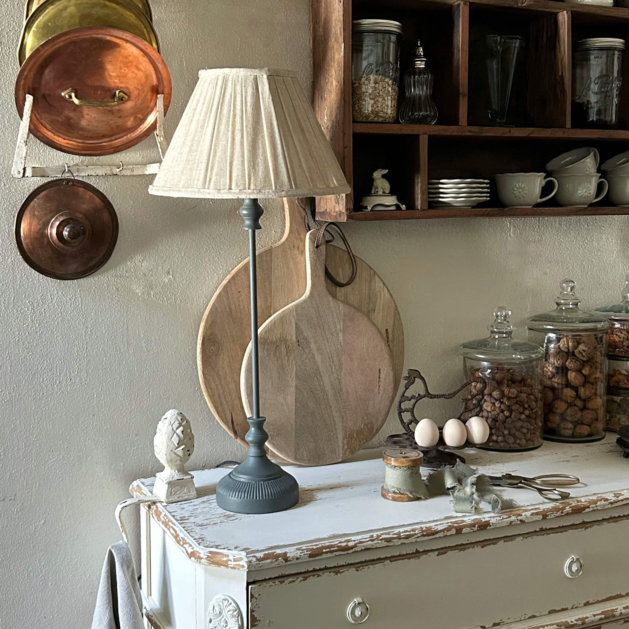 Lámpara de mesa con base gris y pantalla crema sobre cómoda blanca envejecida en cocina rústica
