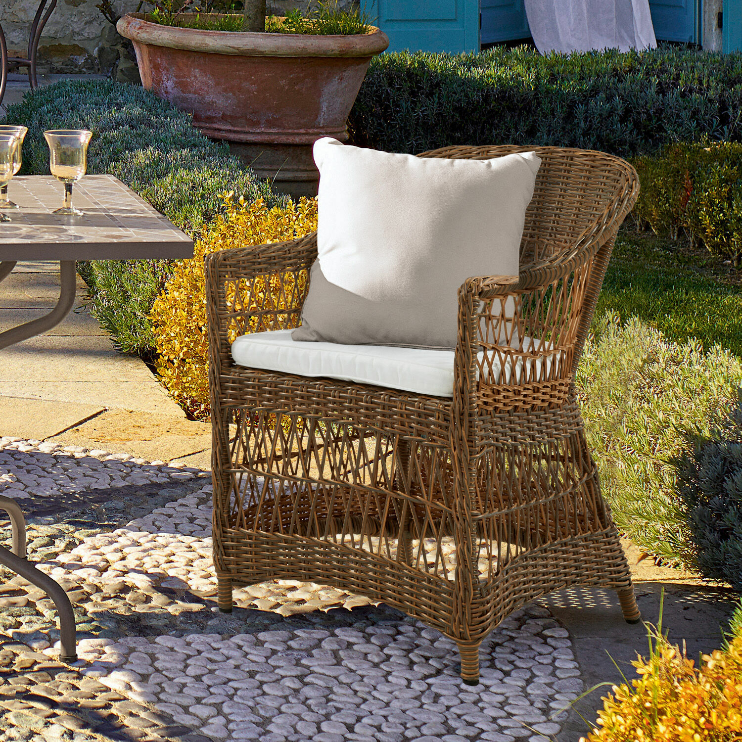 Sillón de ratán con cojín blanco sobre suelo de piedra en jardín soleado para muebles exteriores