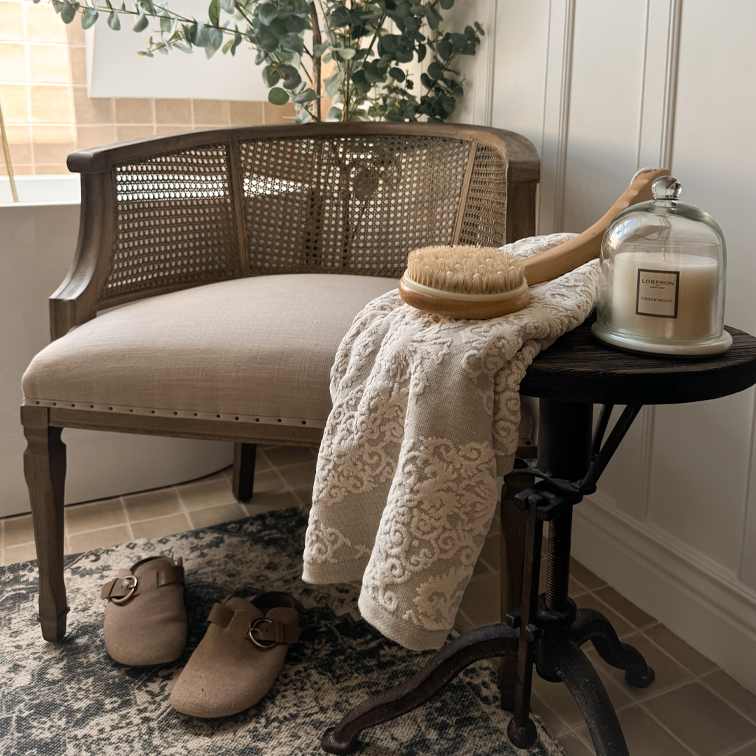 Silla de madera con asiento claro, toalla con patrón, cepillo corporal y vela aromática sobre mesa auxiliar