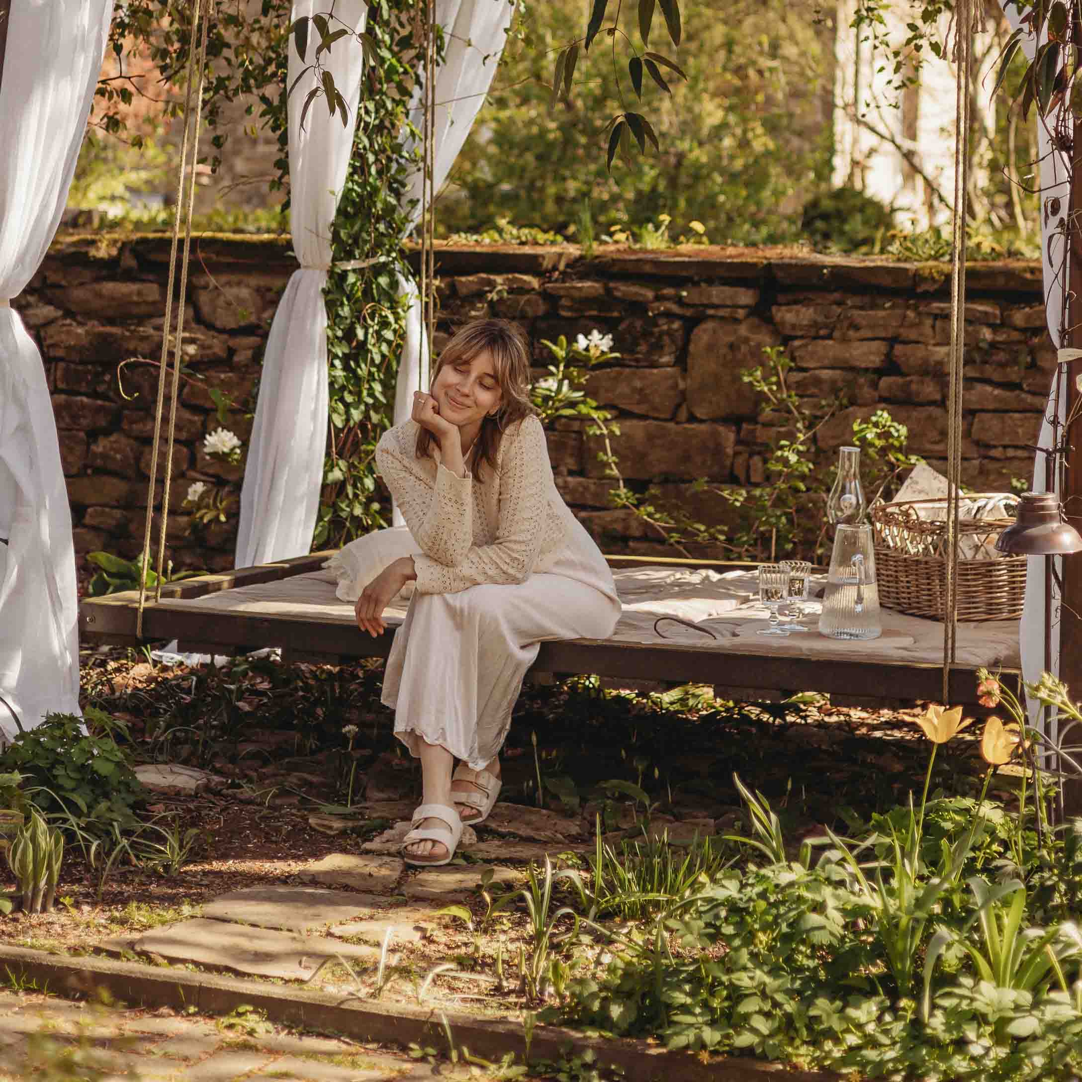Mujer con suéter beige sentada en un columpio de jardín de madera con cortinas blancas y entorno natural