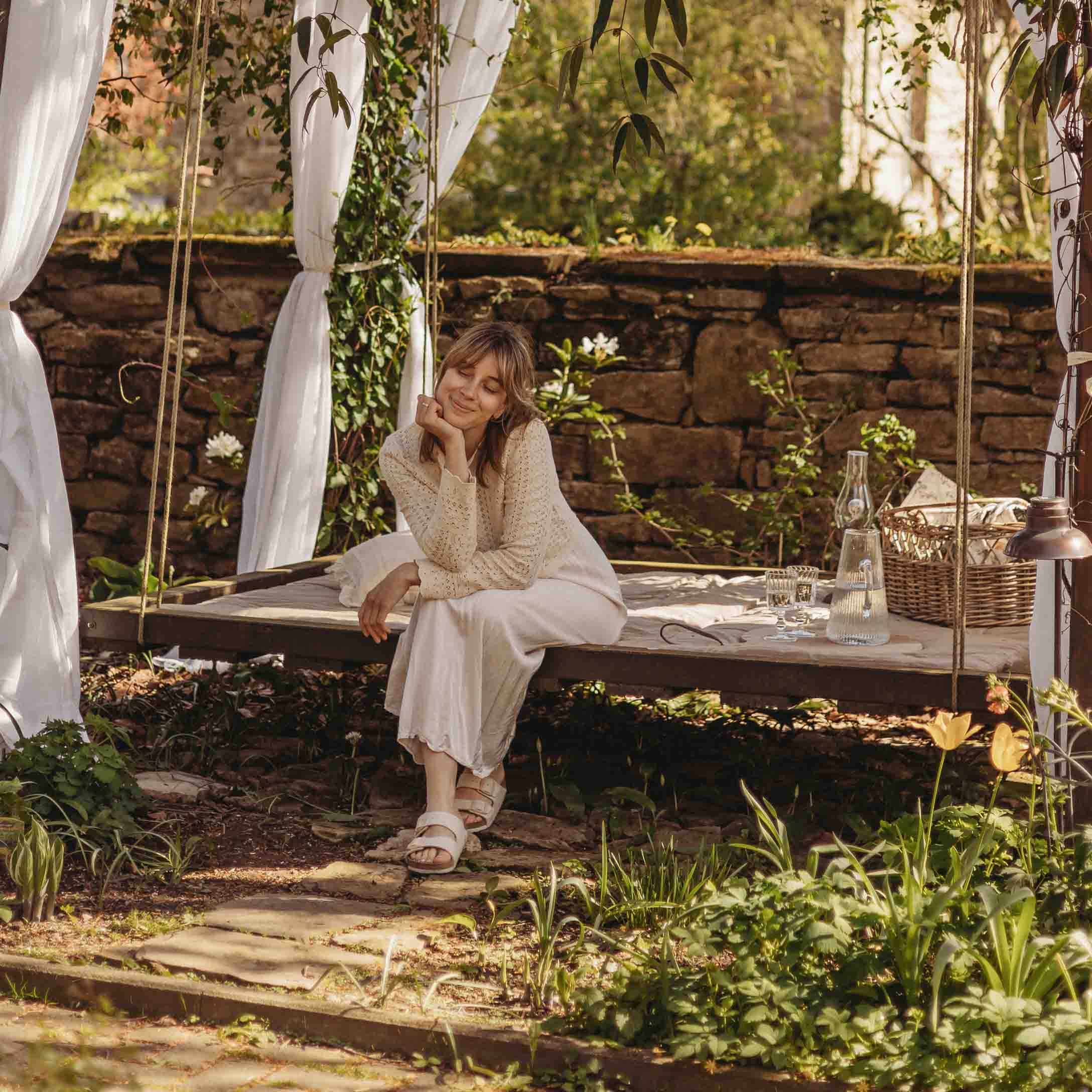 Mujer con suéter beige sentada en un columpio de jardín de madera con cortinas blancas y entorno natural