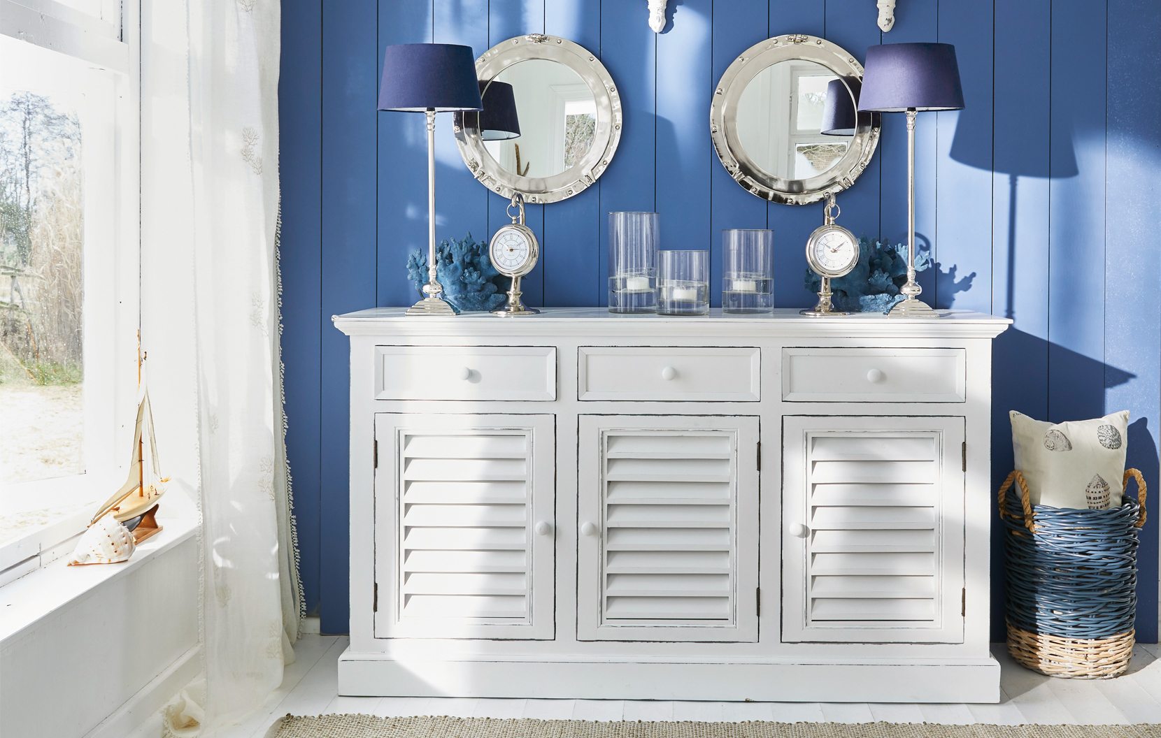 Aparador blanco de madera con puertas de persiana frente a pared azul con decoración marina