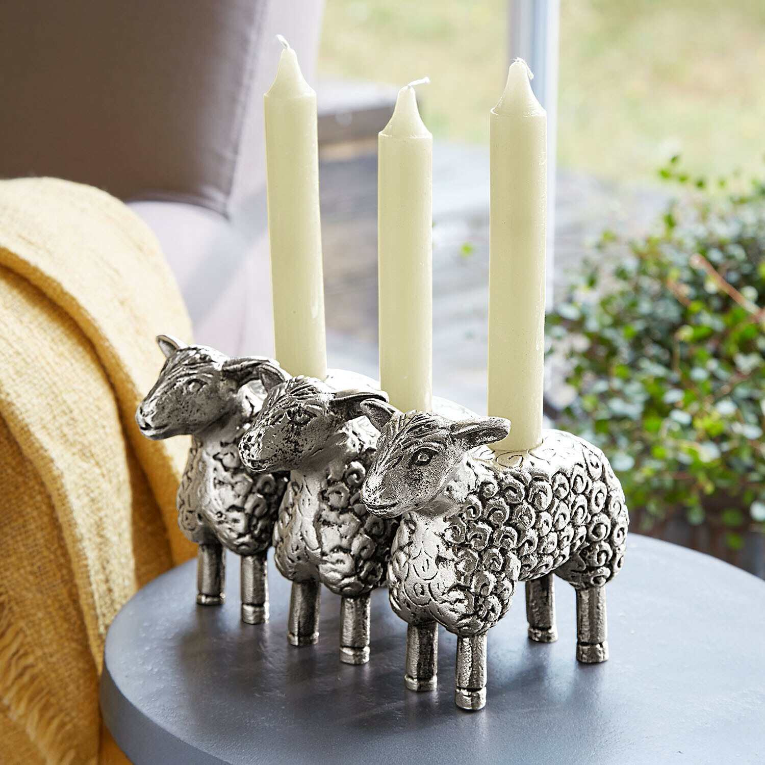 Portavelas Séverac Tres candelabros plateados con forma de oveja y velas blancas sobre mesa gris en sala