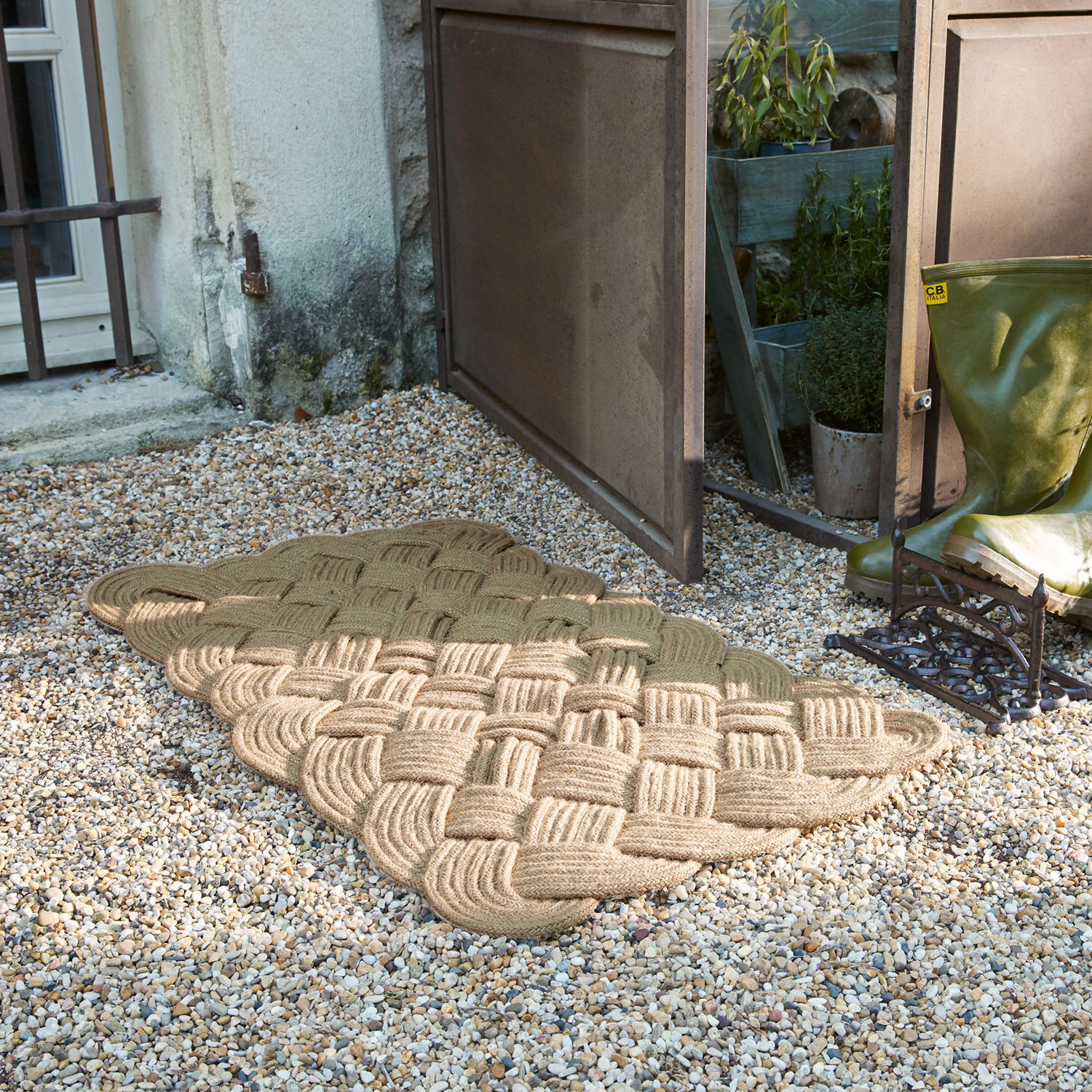Felpudo beige de cuerda trenzada sobre grava junto a puerta abierta y botas de agua verdes