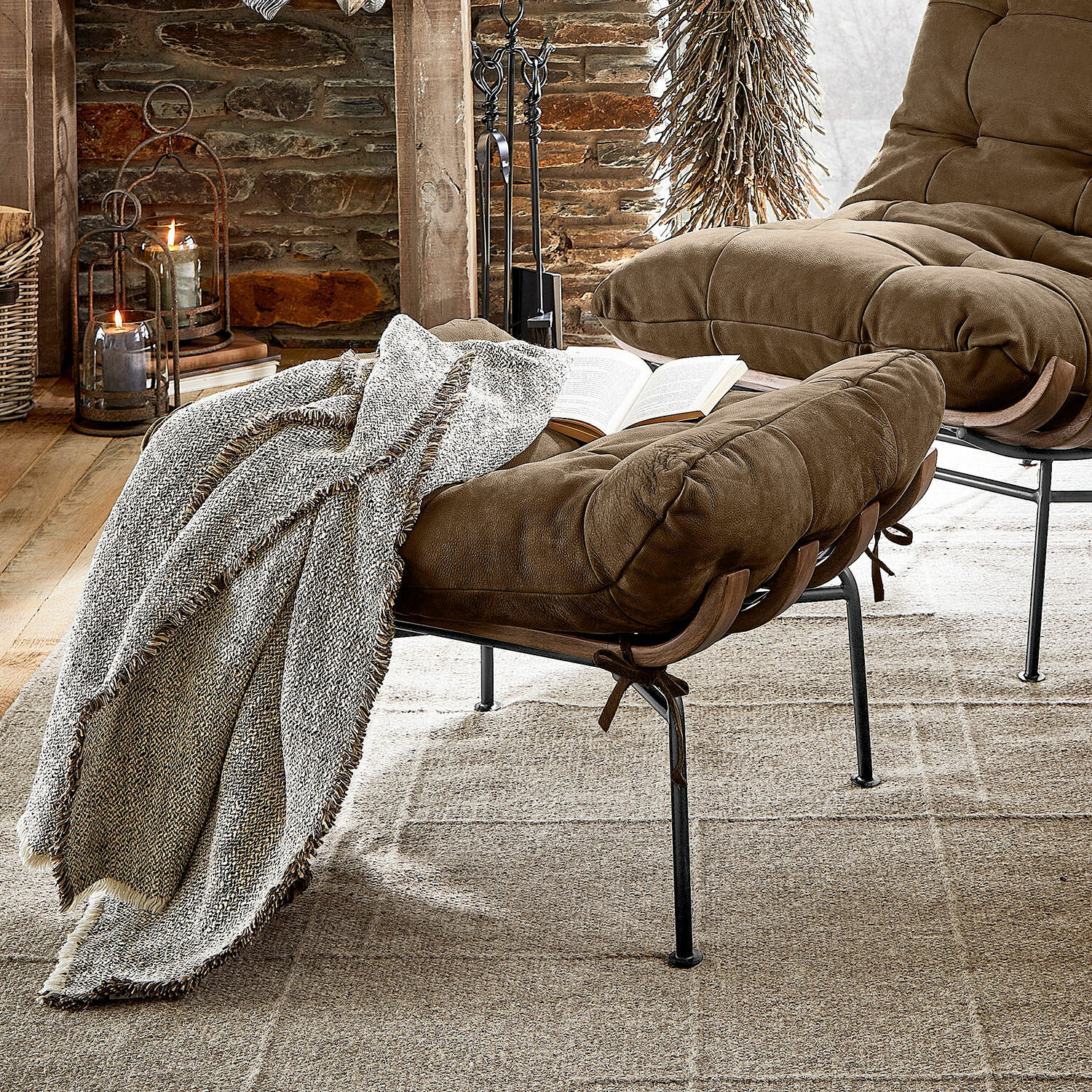 Taburete Horraine Sillón marrón de tela suave con reposapiés y manta gris sobre alfombra beige en interior rústico