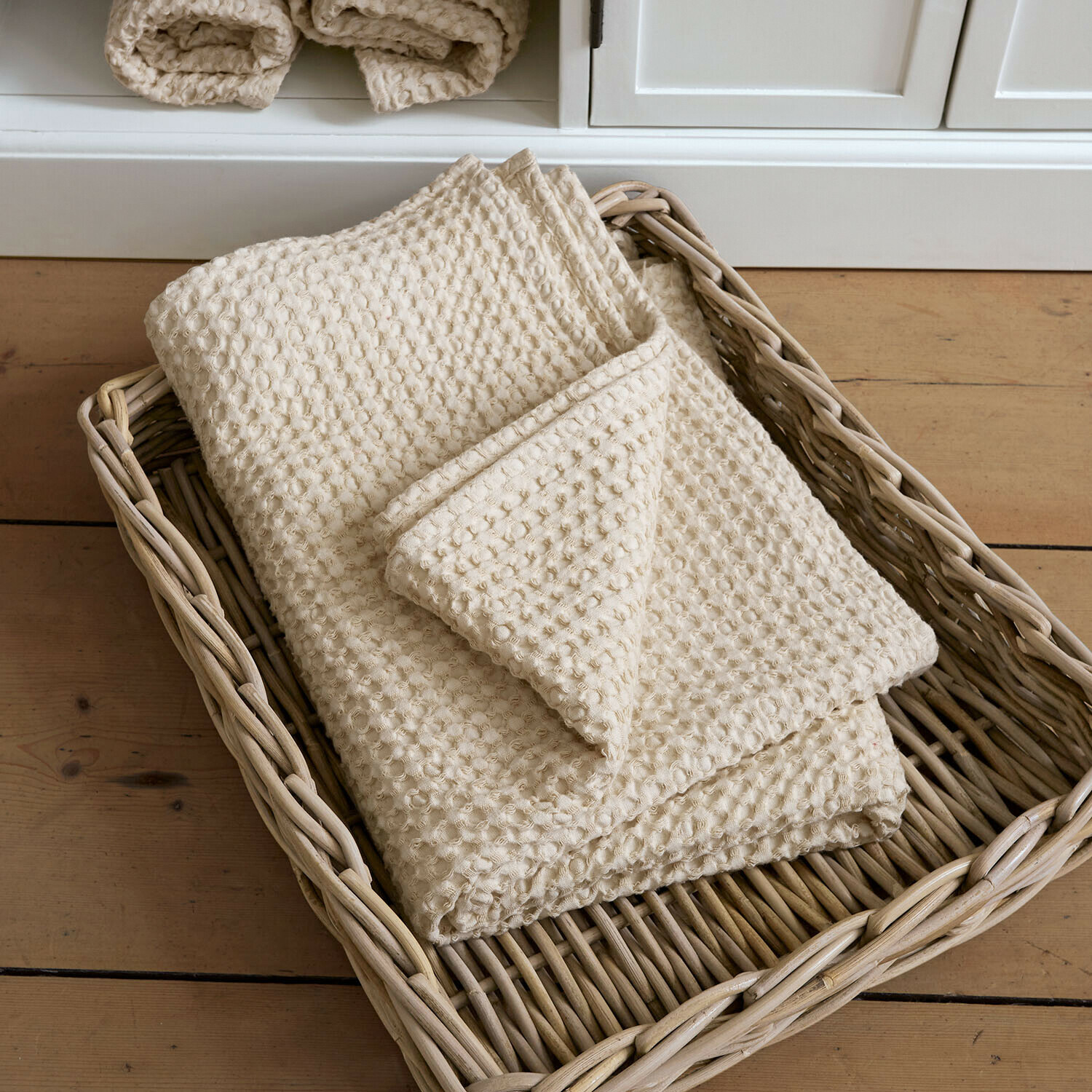 Toalla de baño Maralei Manta de algodón color crema con textura de gofre en cesta de mimbre sobre suelo de madera