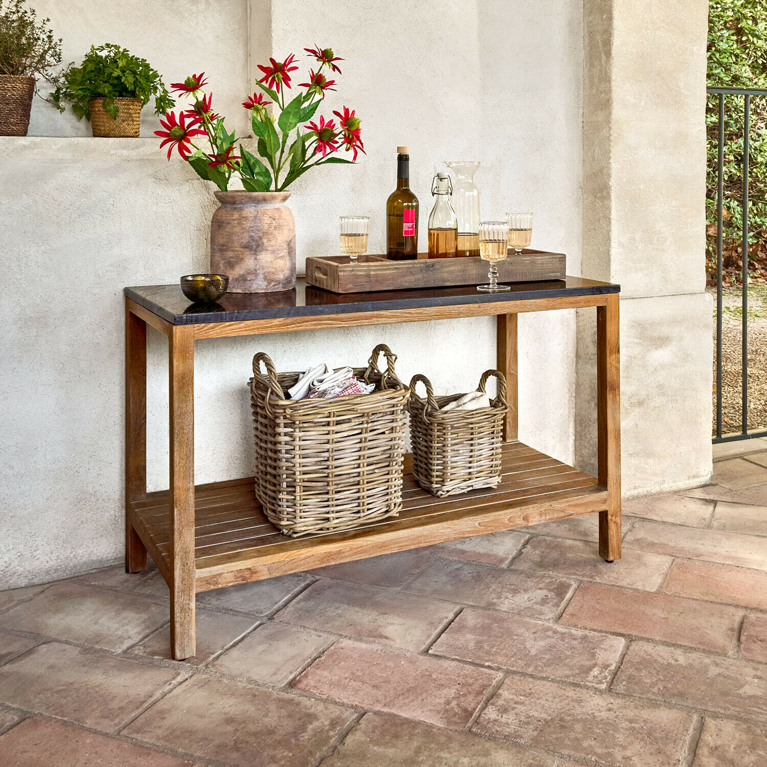 Mesa para cocina exterior Corviano Consola de madera con superficie negra, decorada con jarrón de flores rojas y cestas de mimbre en la repisa inferior