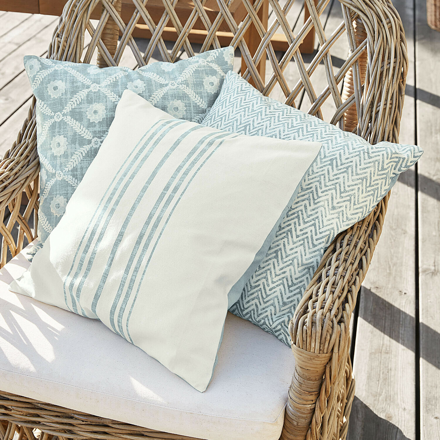 Tres cojines decorativos en tonos azul claro y blanco sobre una silla de mimbre en terraza de madera