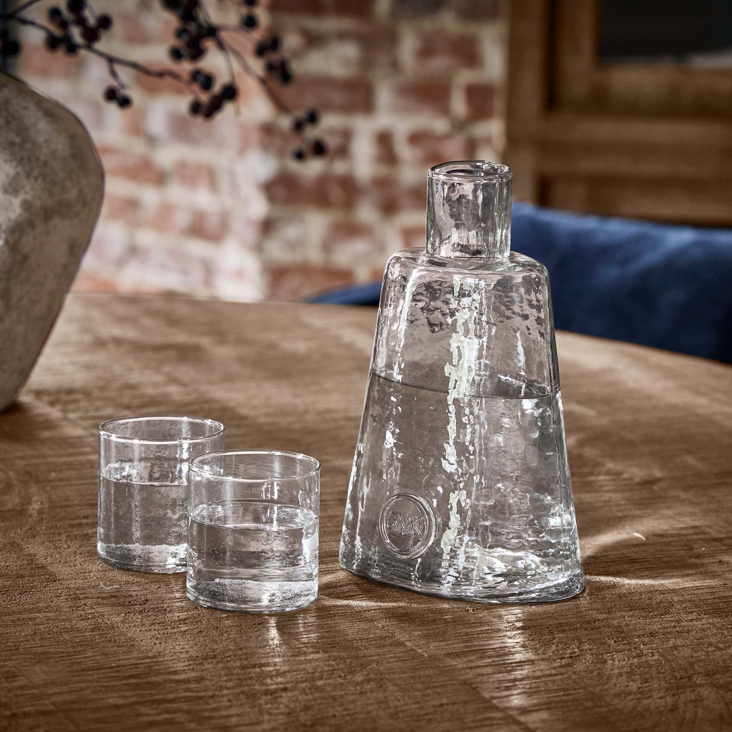 Jarra de vidrio transparente con dos vasos sobre mesa de madera en ambiente rústico